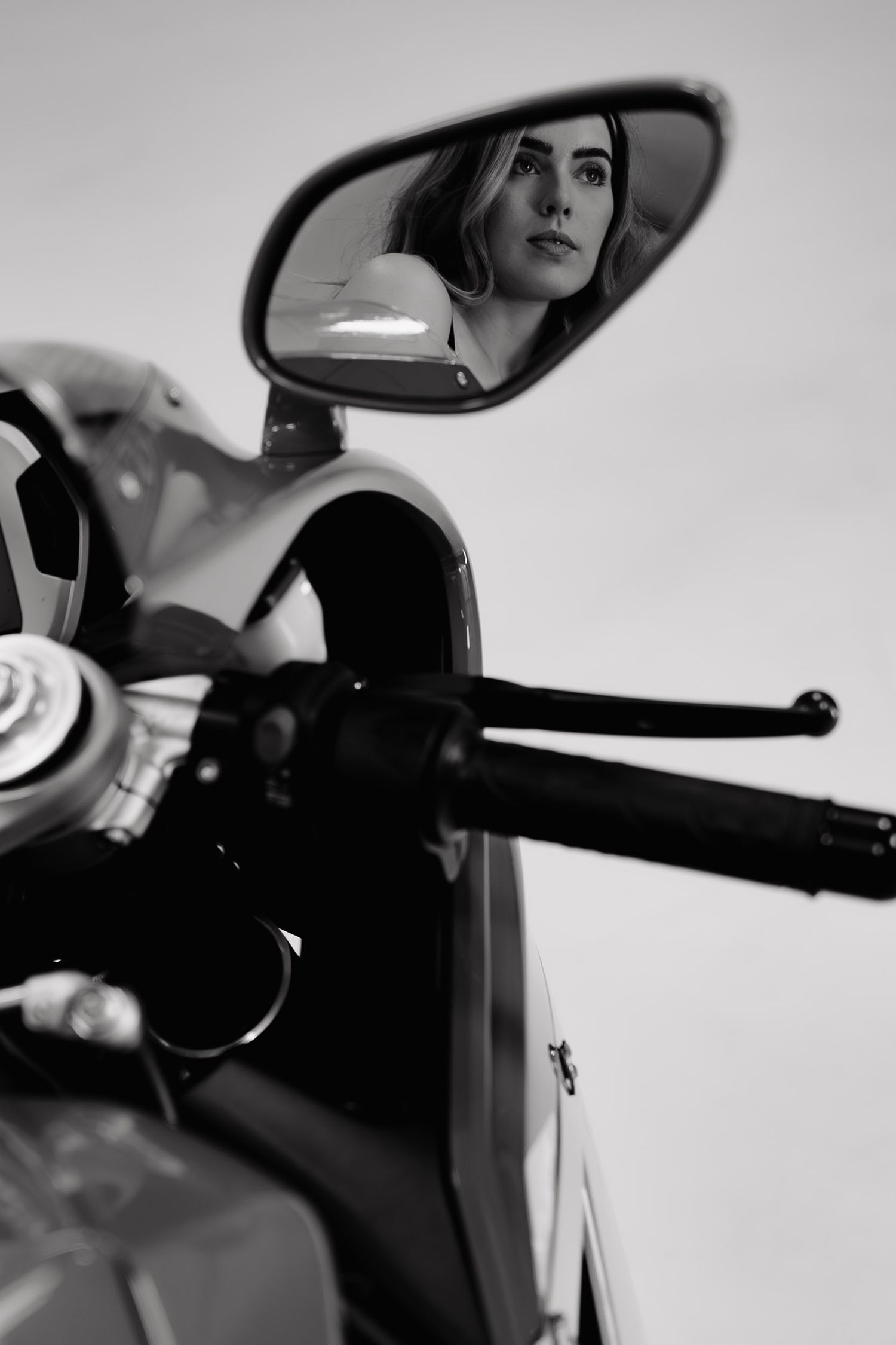 A black and white reflection in a motorcycle side mirror showing someone looking through the mirror while seated on a bike.