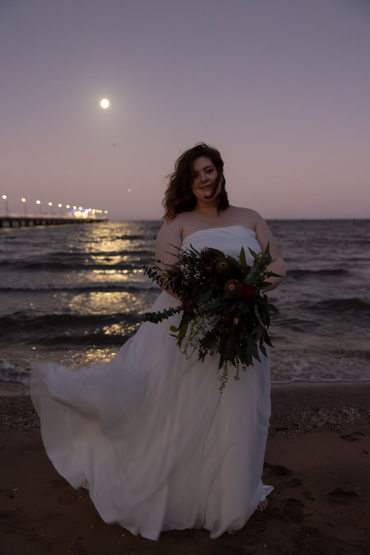 A bride in a flowing white dress holds a greenery bouquet on a beach at dusk with a pier and moonlit water in the background.
