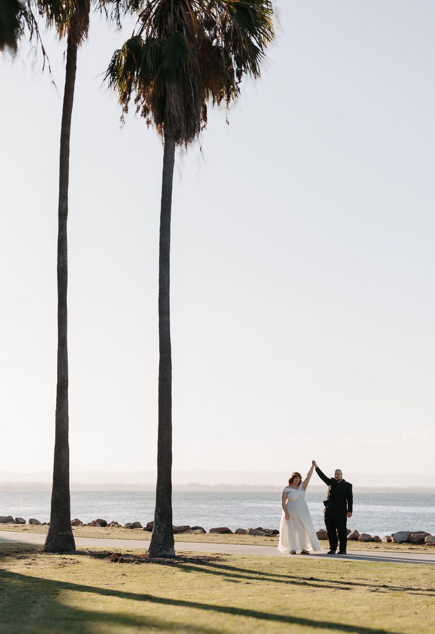 Two silhouettes dance under tall palm trees by the ocean on a sunny day at a waterfront beach wedding ceremony.