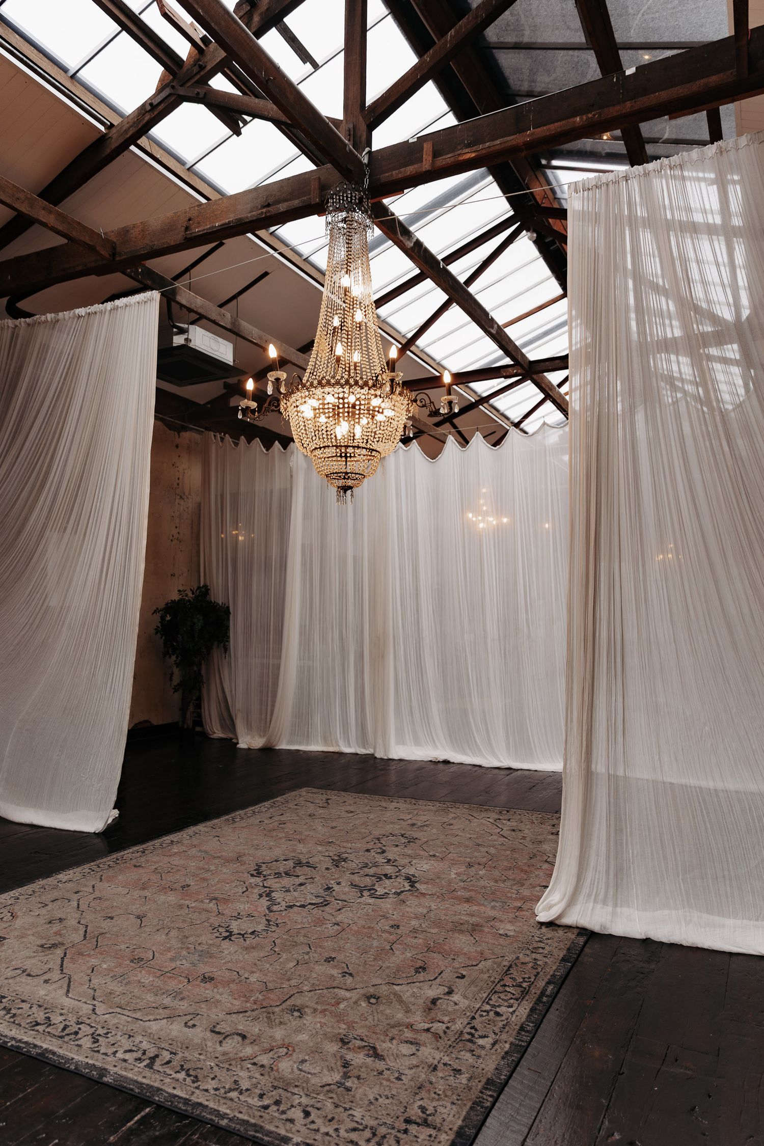 Elegant wedding venue setup with crystal chandelier, flowing white drapes, and vintage rug under exposed wooden beams.