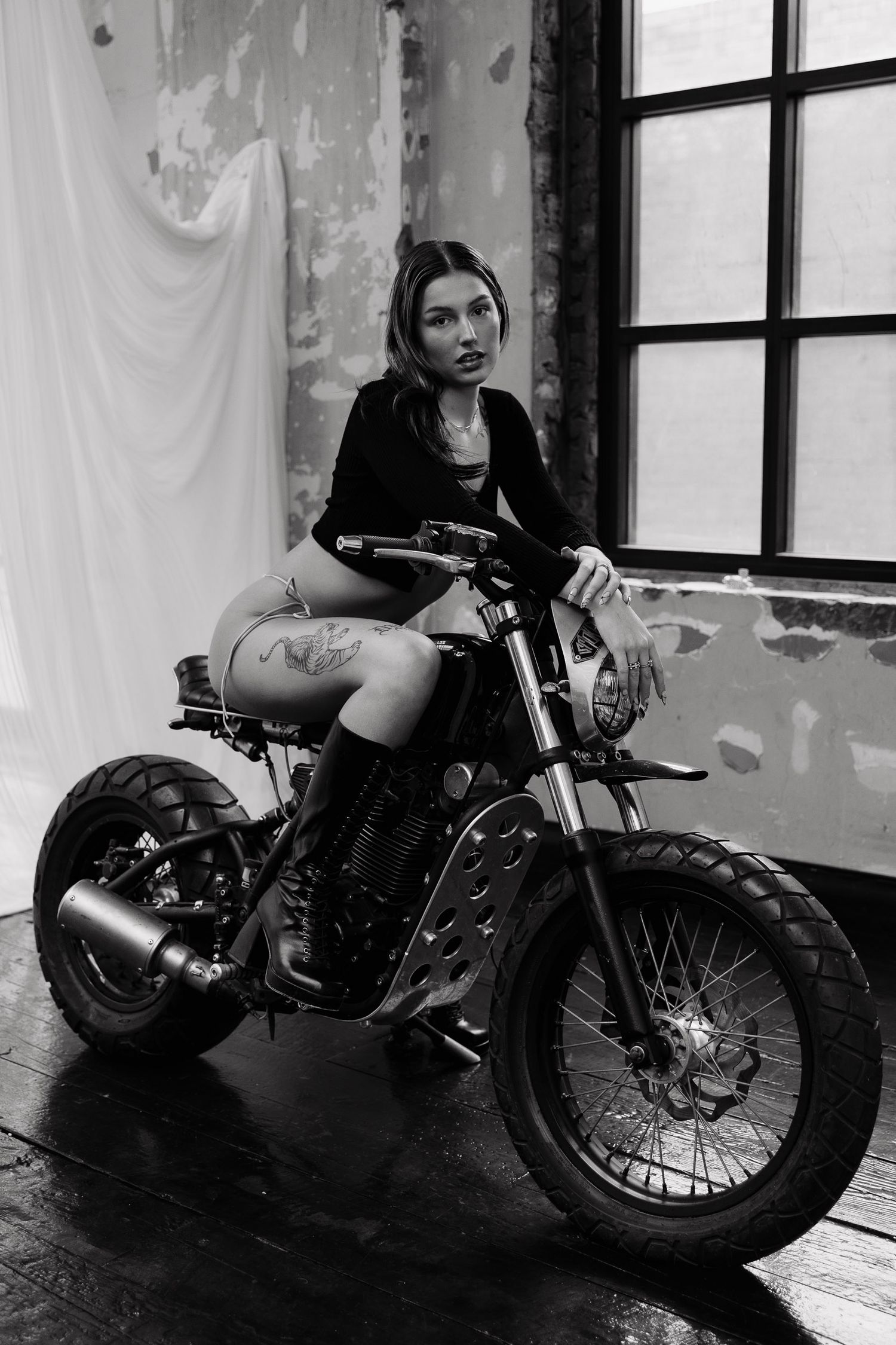 Black and white photo of person sitting on a custom motorcycle in an industrial indoor setting with white draped backdrop.