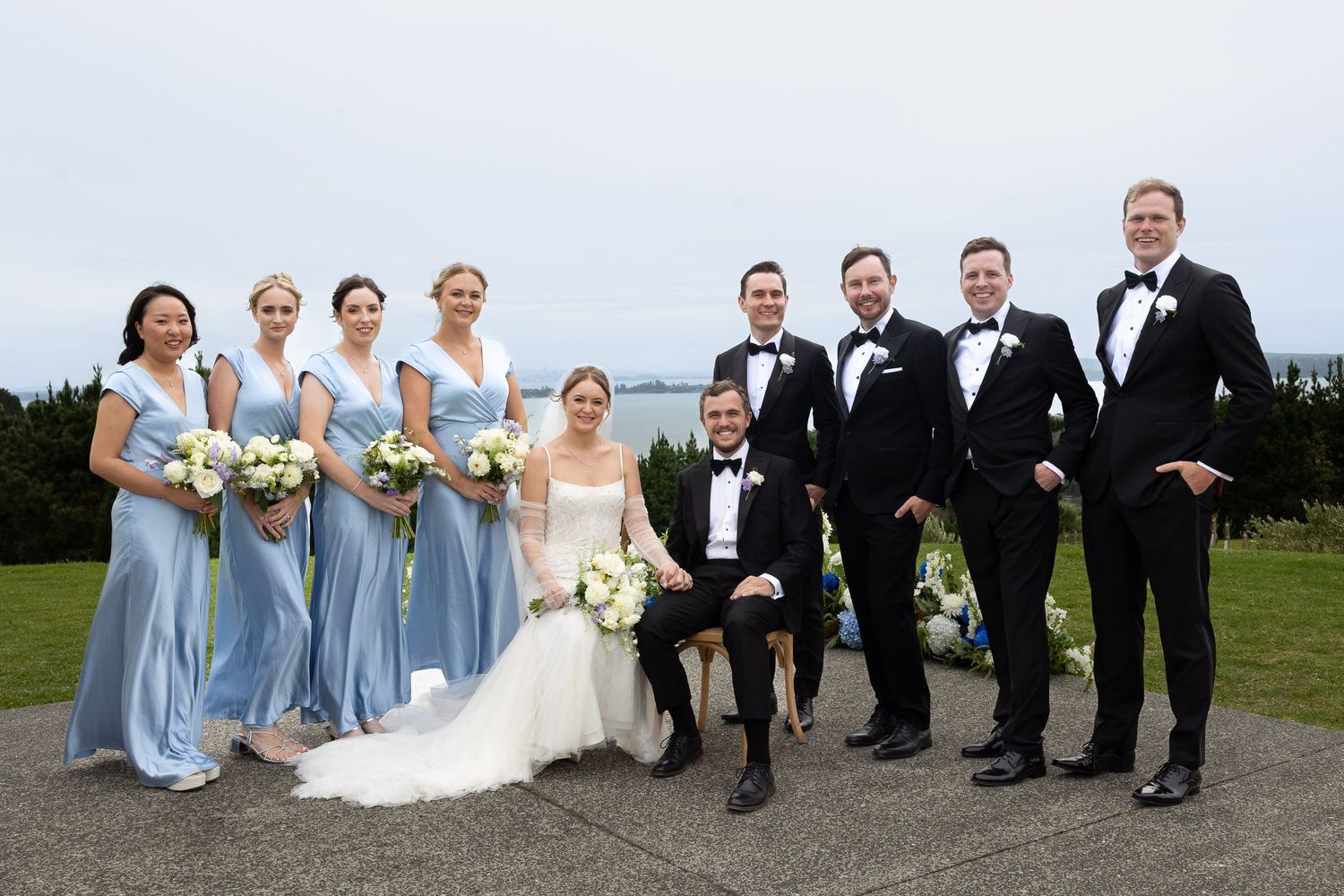 Wedding party in light blue bridesmaid dresses and black tuxedos pose together outdoors on overcast day.