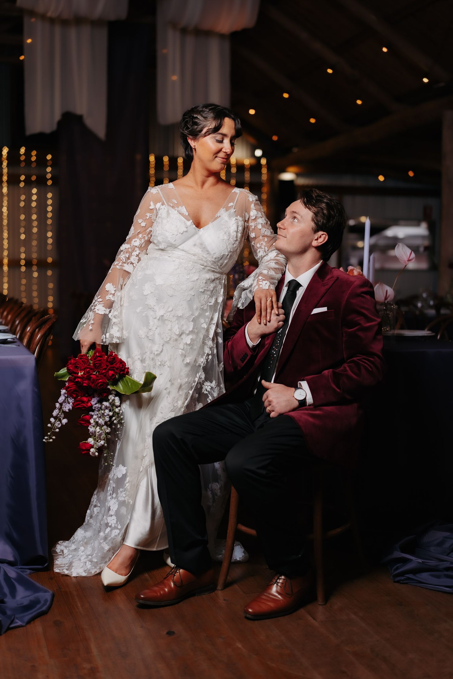 Wedding couple shares intimate moment during garter removal ceremony on dimly lit dance floor with string lights above.