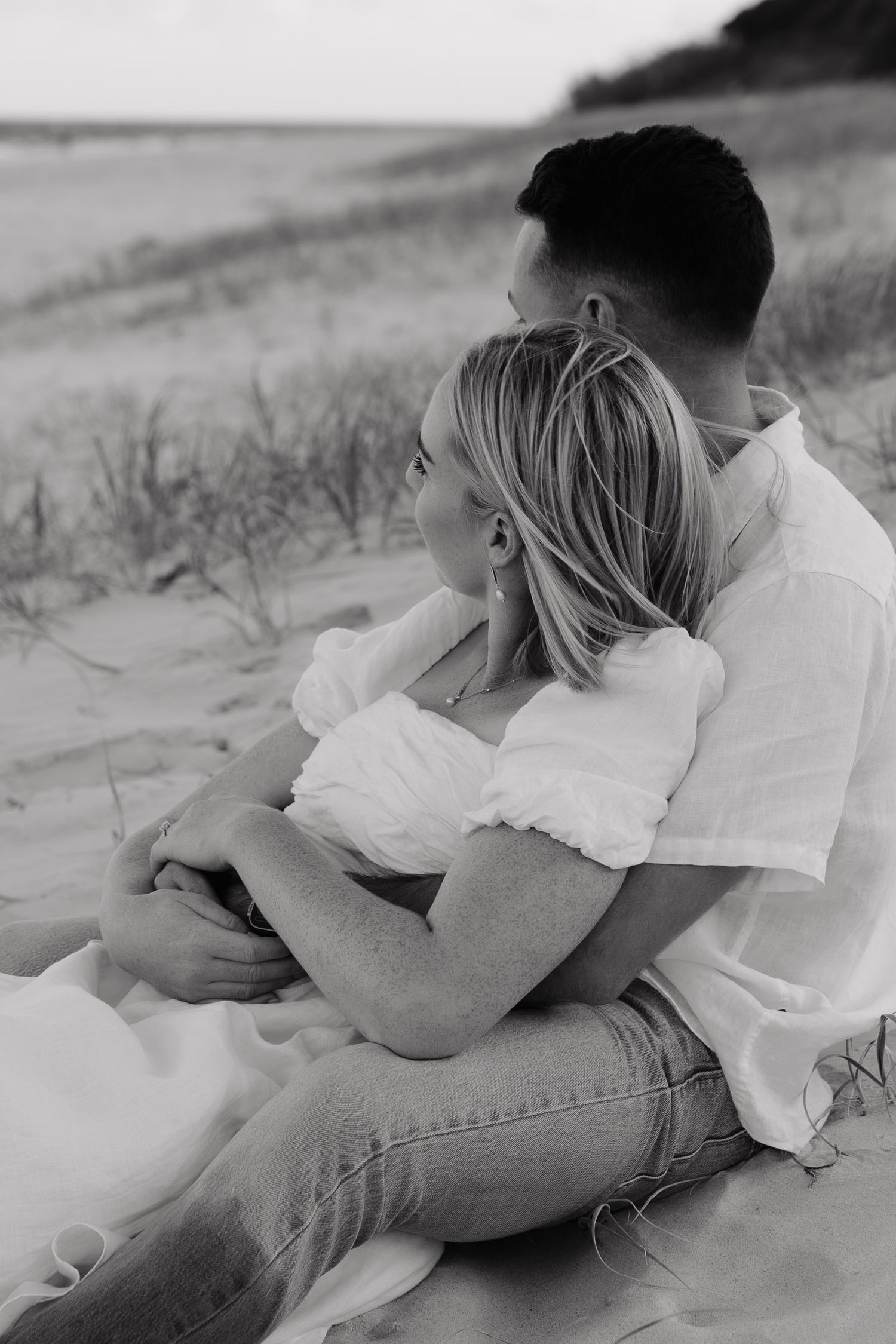 A romantic black and white photo of a couple embracing on a sandy beach while sitting together at sunset.