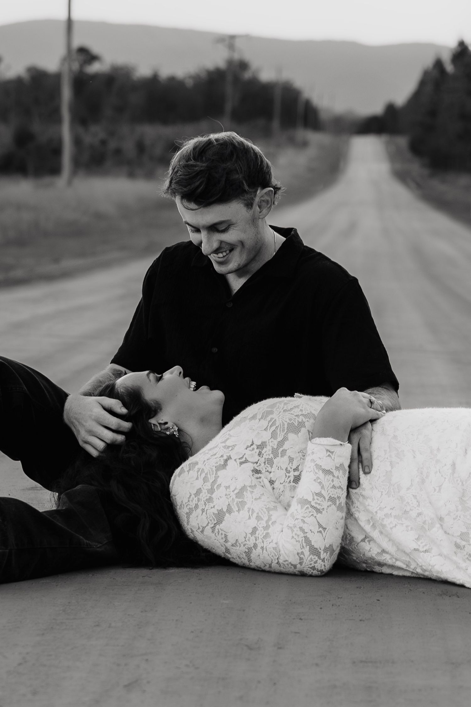 A tender black and white romantic moment between two people laying on an empty rural road.