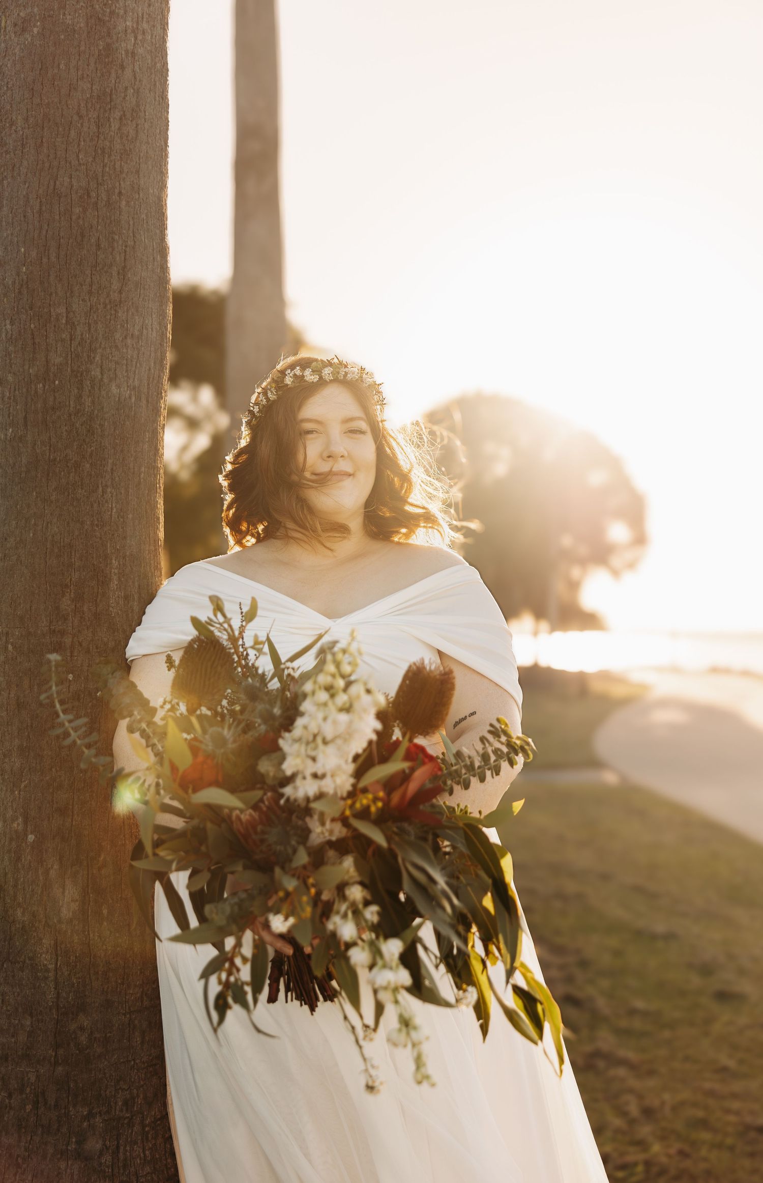 A bride in an off-shoulder white gown holds a lush wildflower bouquet while standing beside a tree during sunset.