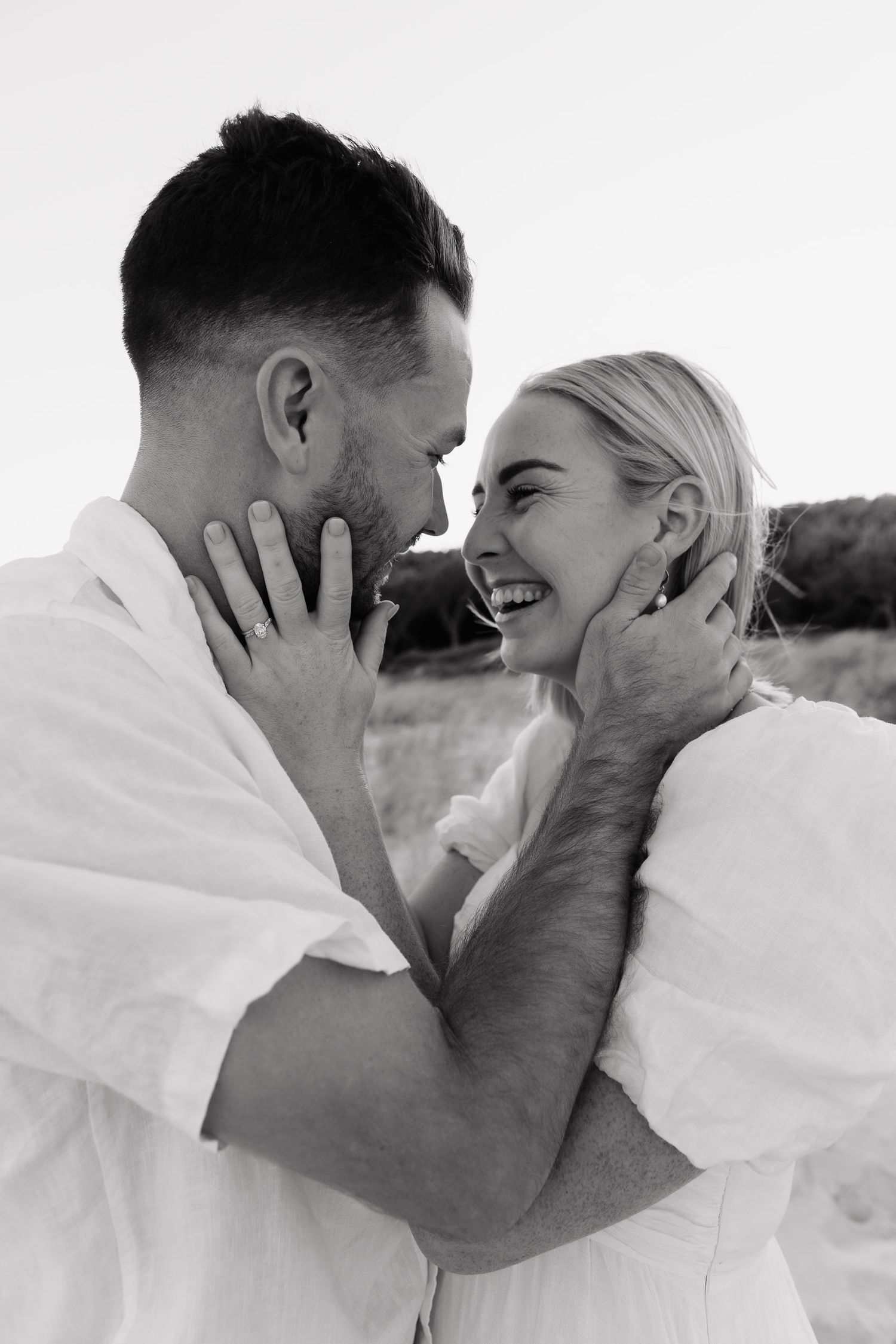 A romantic black and white portrait captures an intimate moment between a couple sharing laughter outdoors.