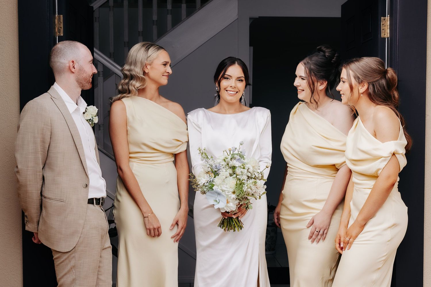 Wedding party members in cream and yellow dresses pose together against a dark wall with bouquets.