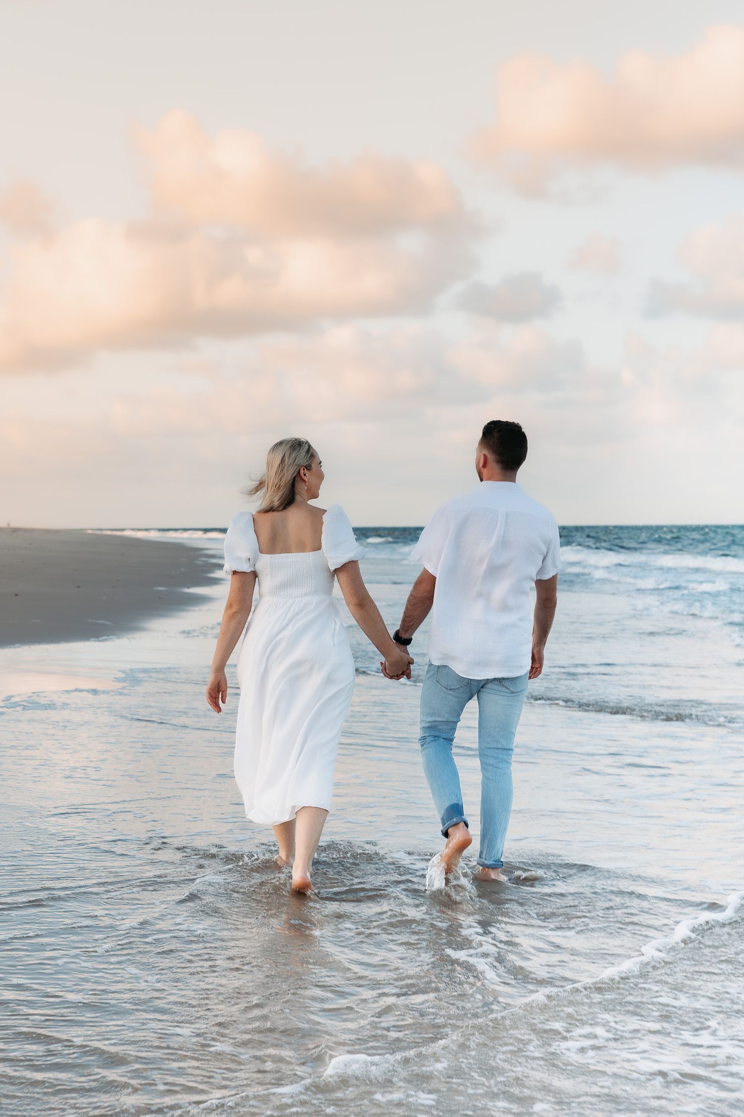 A couple walking hand in hand along the beach at sunset wearing white and casual clothing.