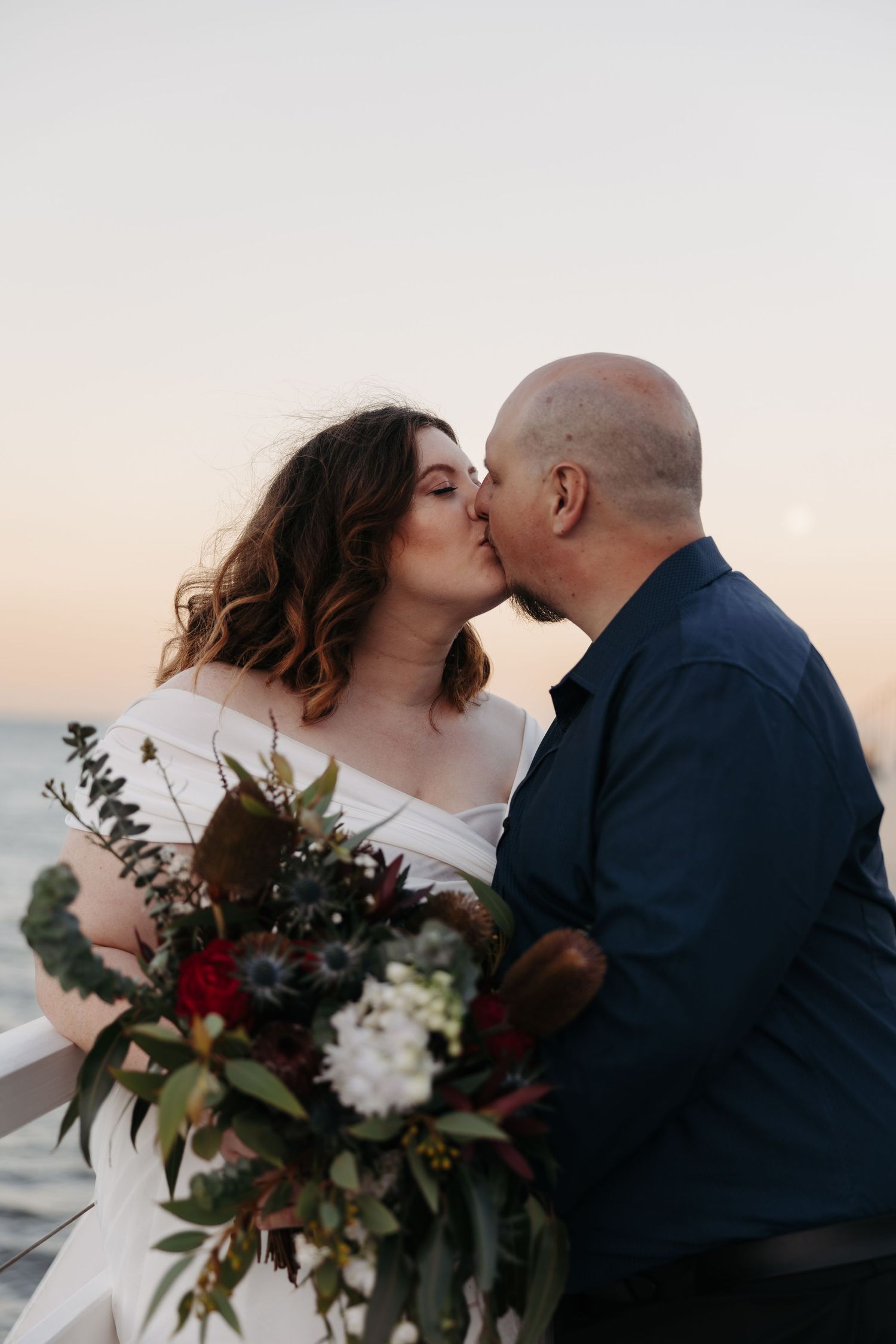 A romantic couple shares a kiss at sunset while holding a bouquet with burgundy and white florals.