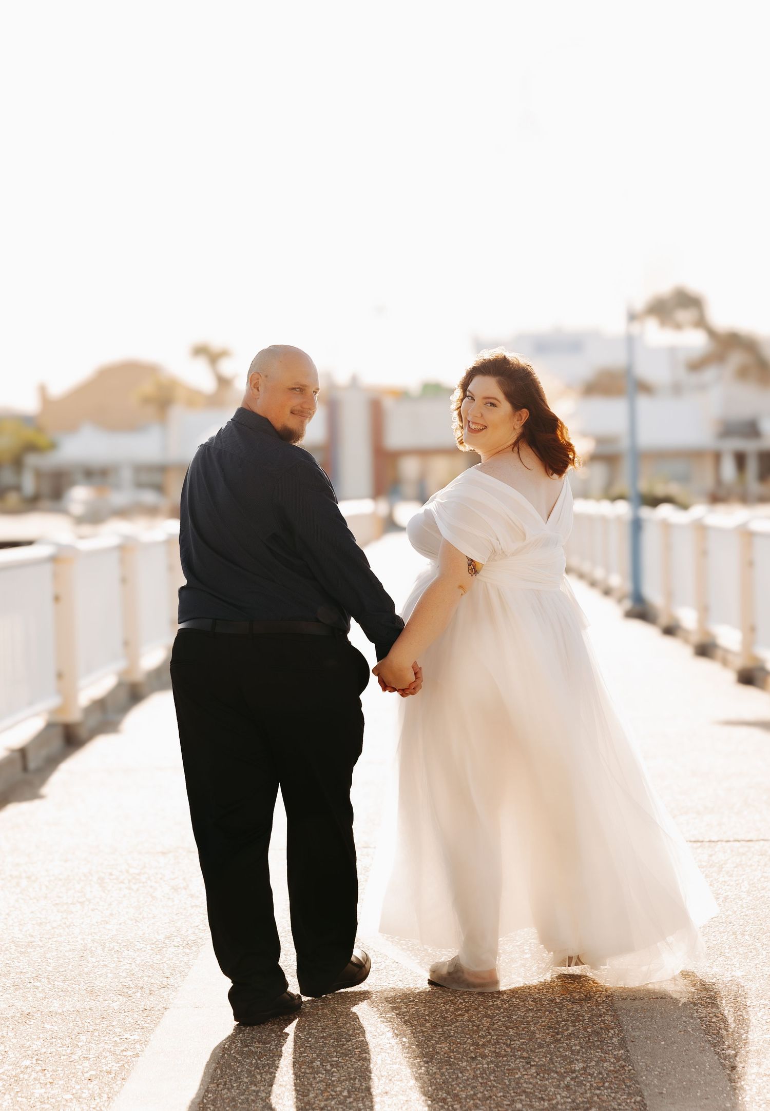 A couple on their wedding day sharing a tender moment on a sunny boardwalk, with the bride in a flowing white gown.