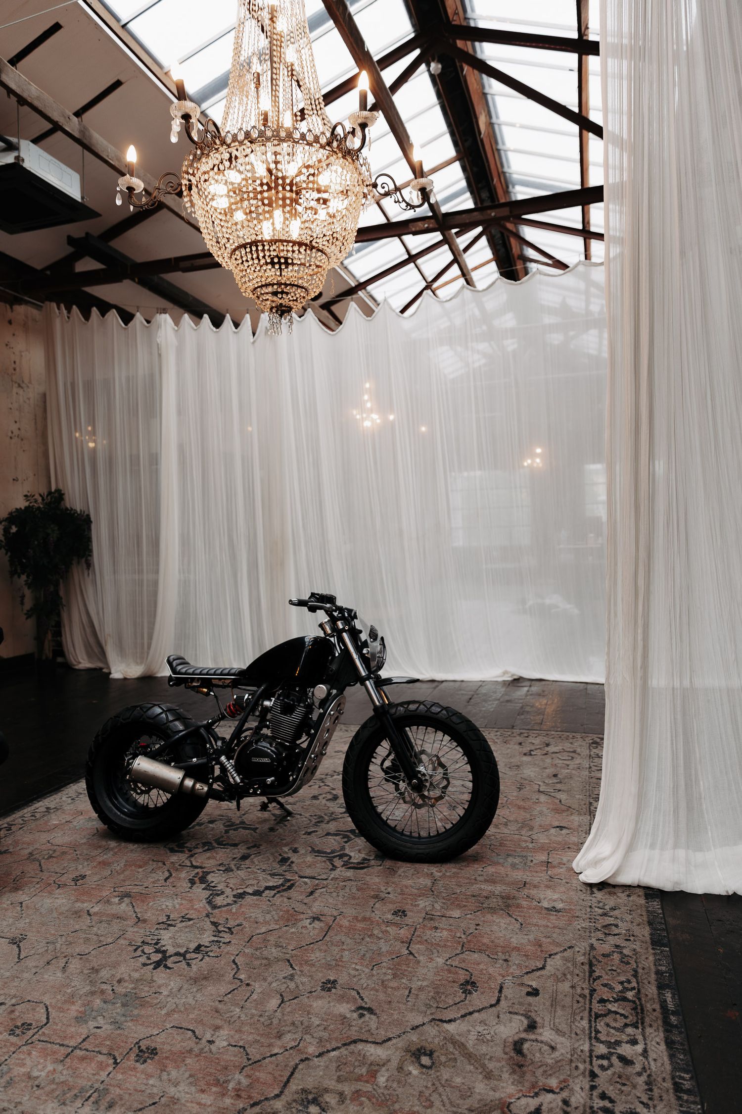 Custom motorcycle displayed under an elegant crystal chandelier with white drapes and exposed beams in an industrial space.