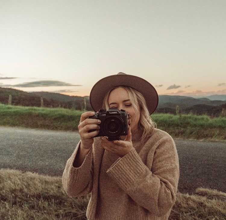 Photographer wearing brown hat and sweater capturing shots with a vintage camera in natural outdoor setting at dusk.