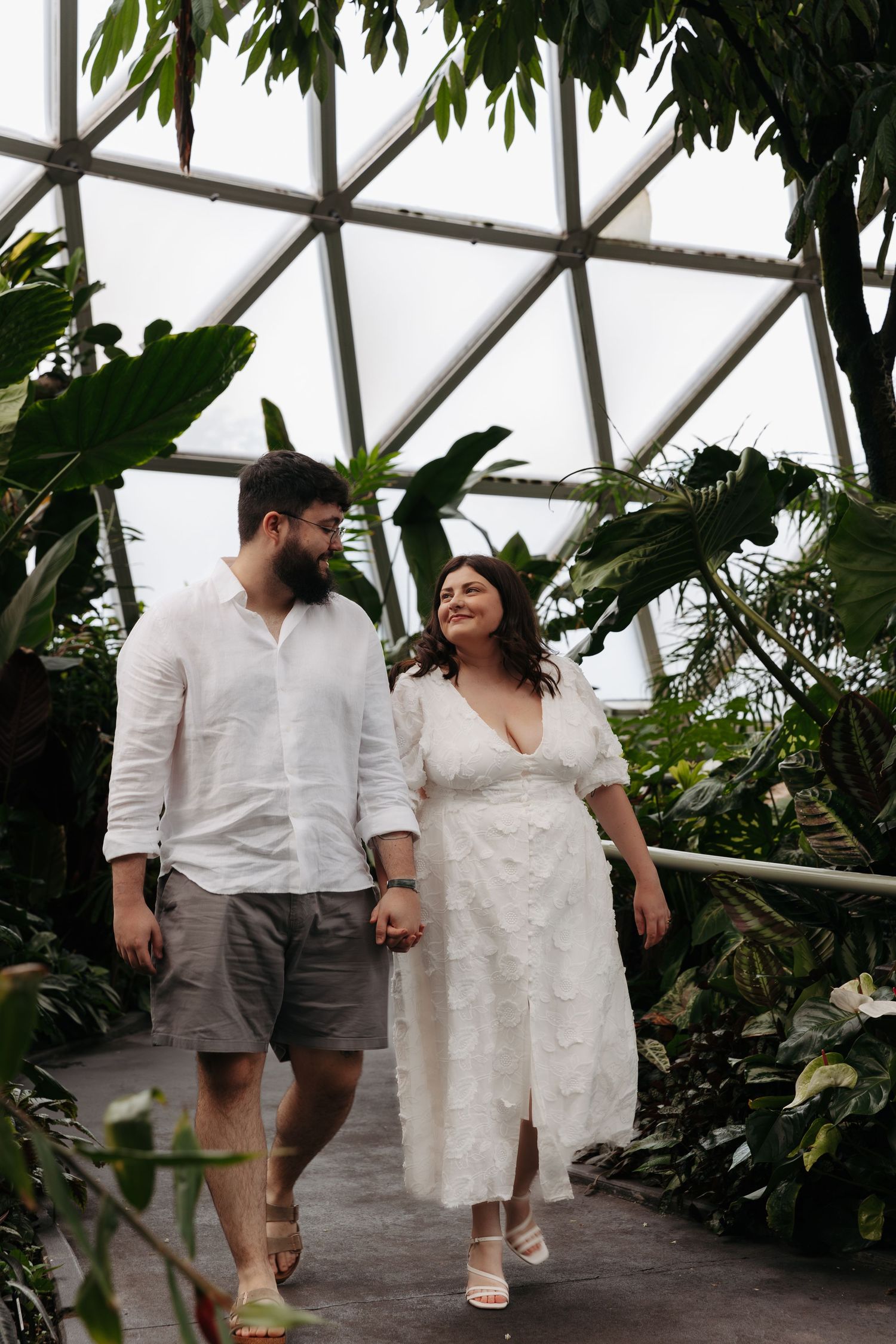 A couple walks hand in hand through a greenhouse filled with lush tropical plants and greenery.