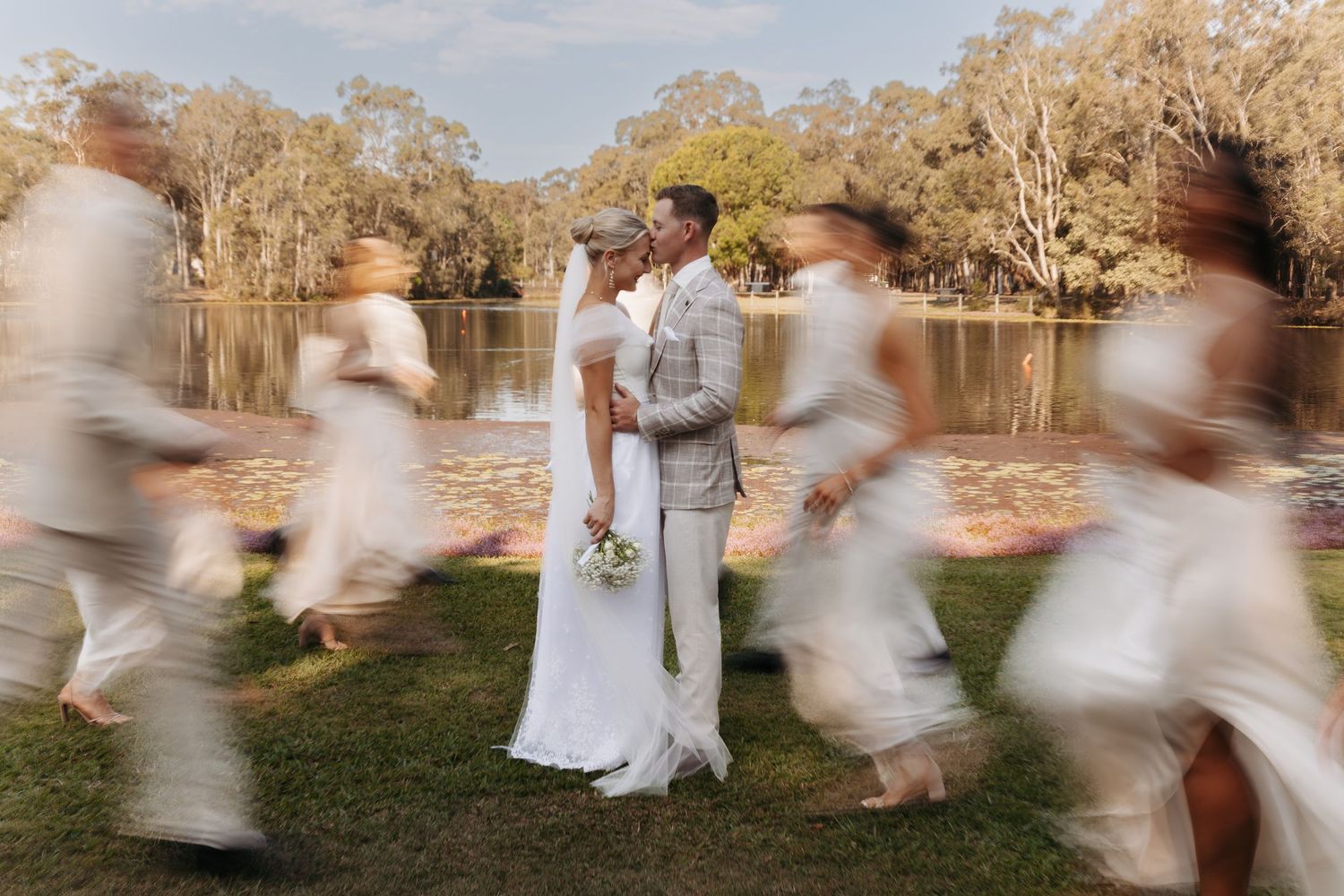 Motion blur captures wedding party running through field with white dresses near lakeside at sunset.