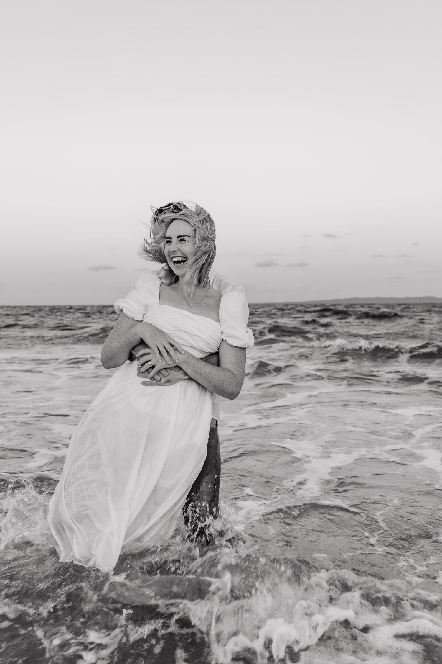 A joyful woman in a white dress stands in ocean waves at the beach in black and white photography.