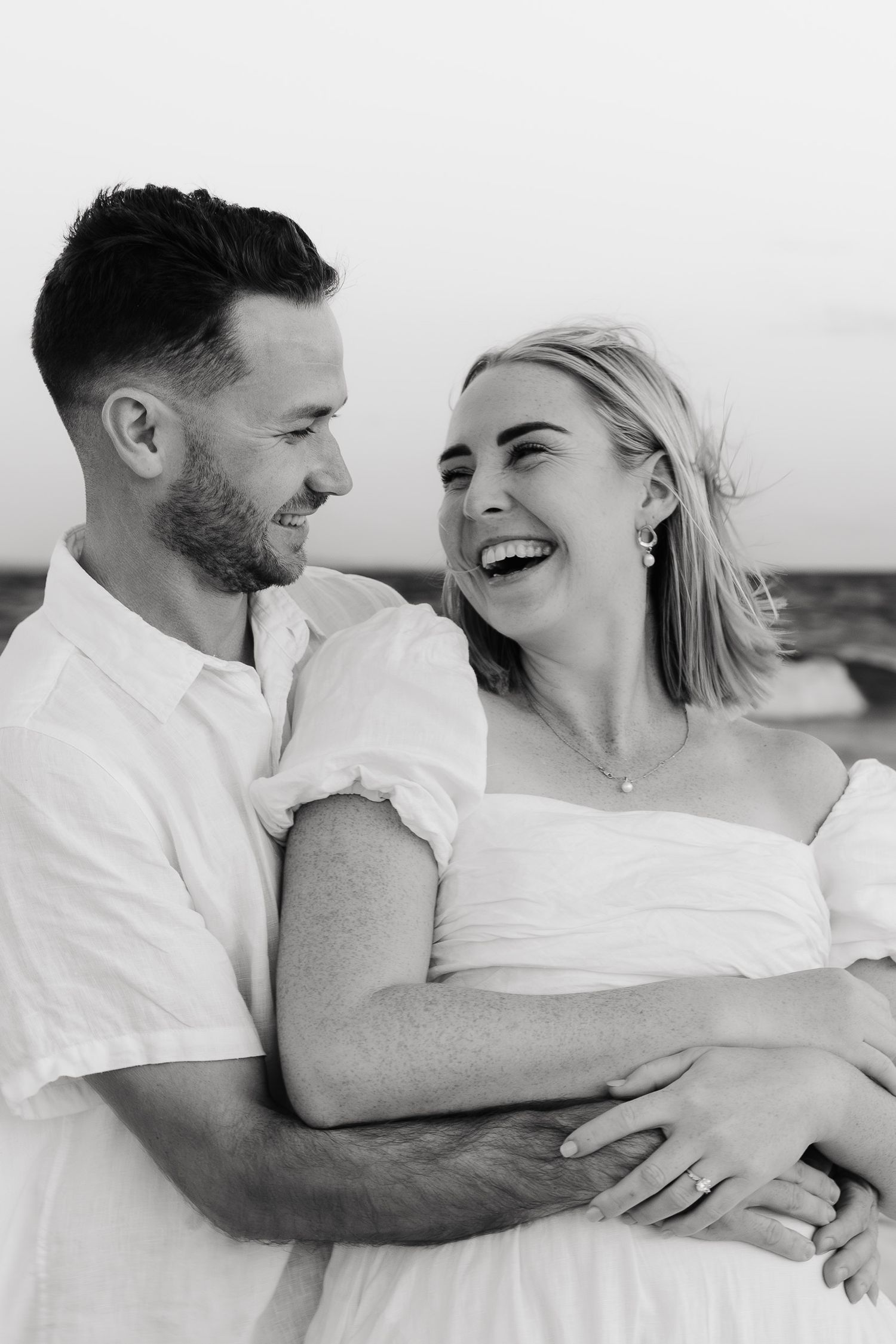 A joyful black and white portrait of a couple embracing and laughing together, both wearing white clothing outdoors.