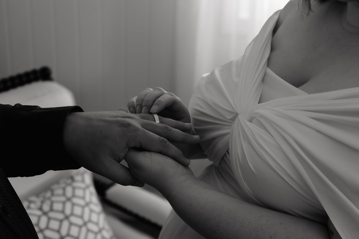 Black and white close-up photograph of hands resting on fabric, showing intimate wedding preparation details.