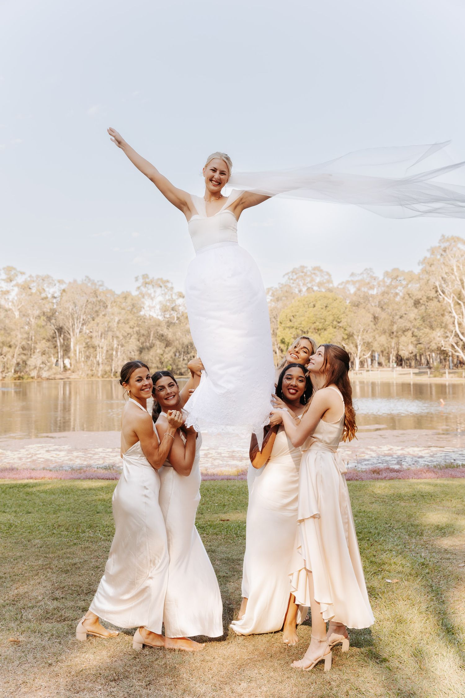 Wedding party in white dresses poses playfully by a lake with bridesmaids lifting the bride in celebration.