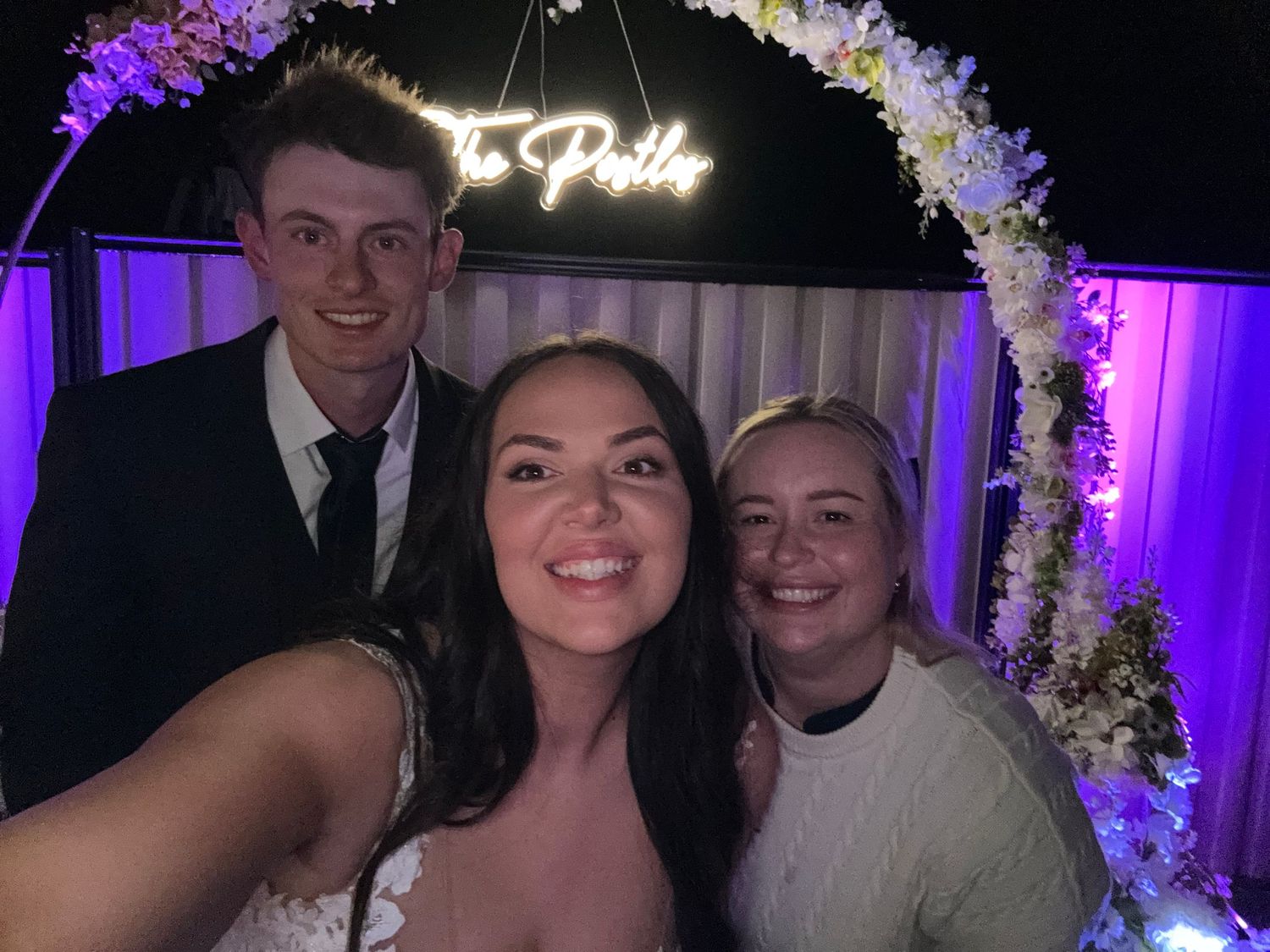 Celebrating guests pose together under a floral arch with purple lighting at an elegant party or wedding event.