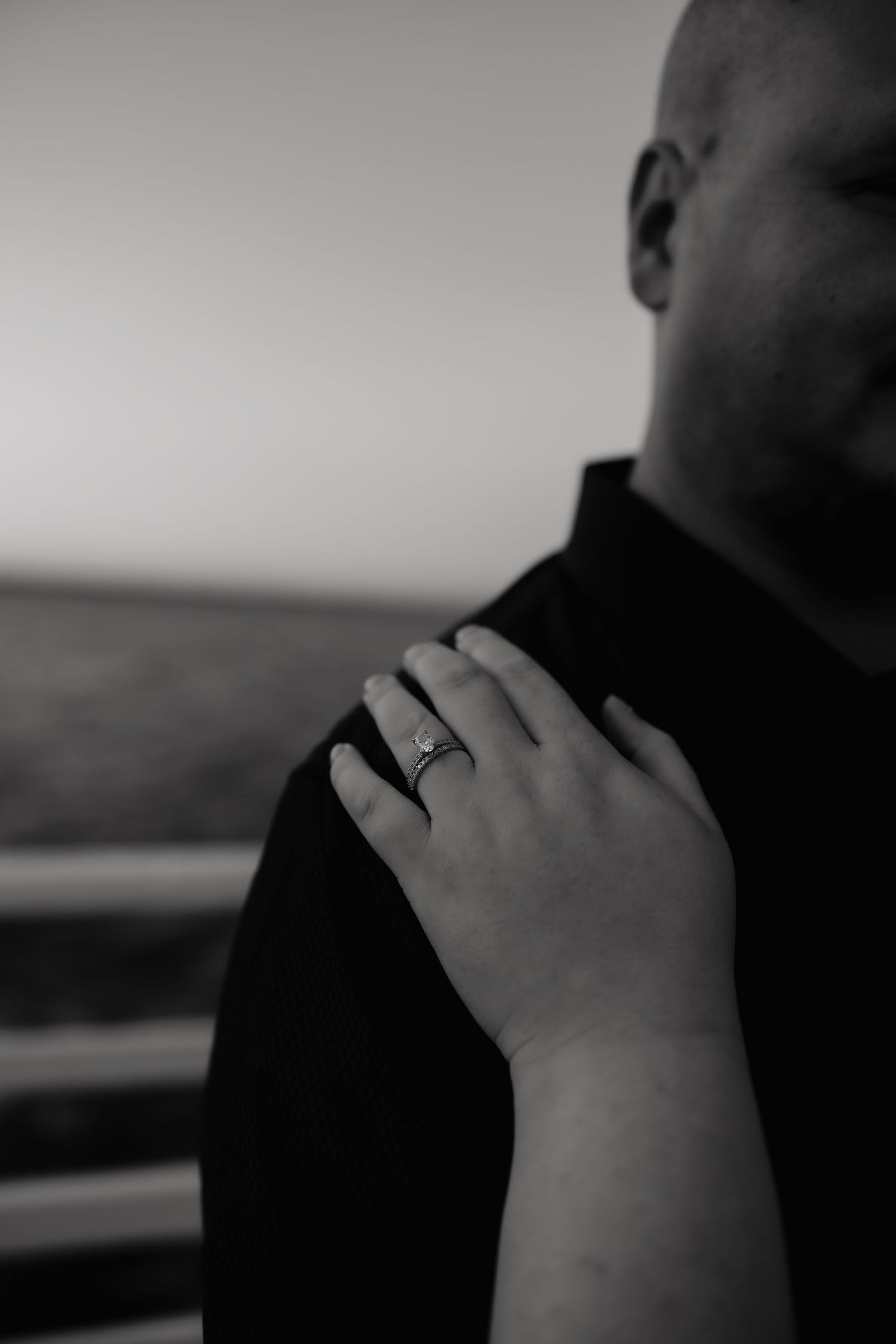 Black and white close-up of a hand wearing a ring resting on someone's shoulder wearing dark clothing.