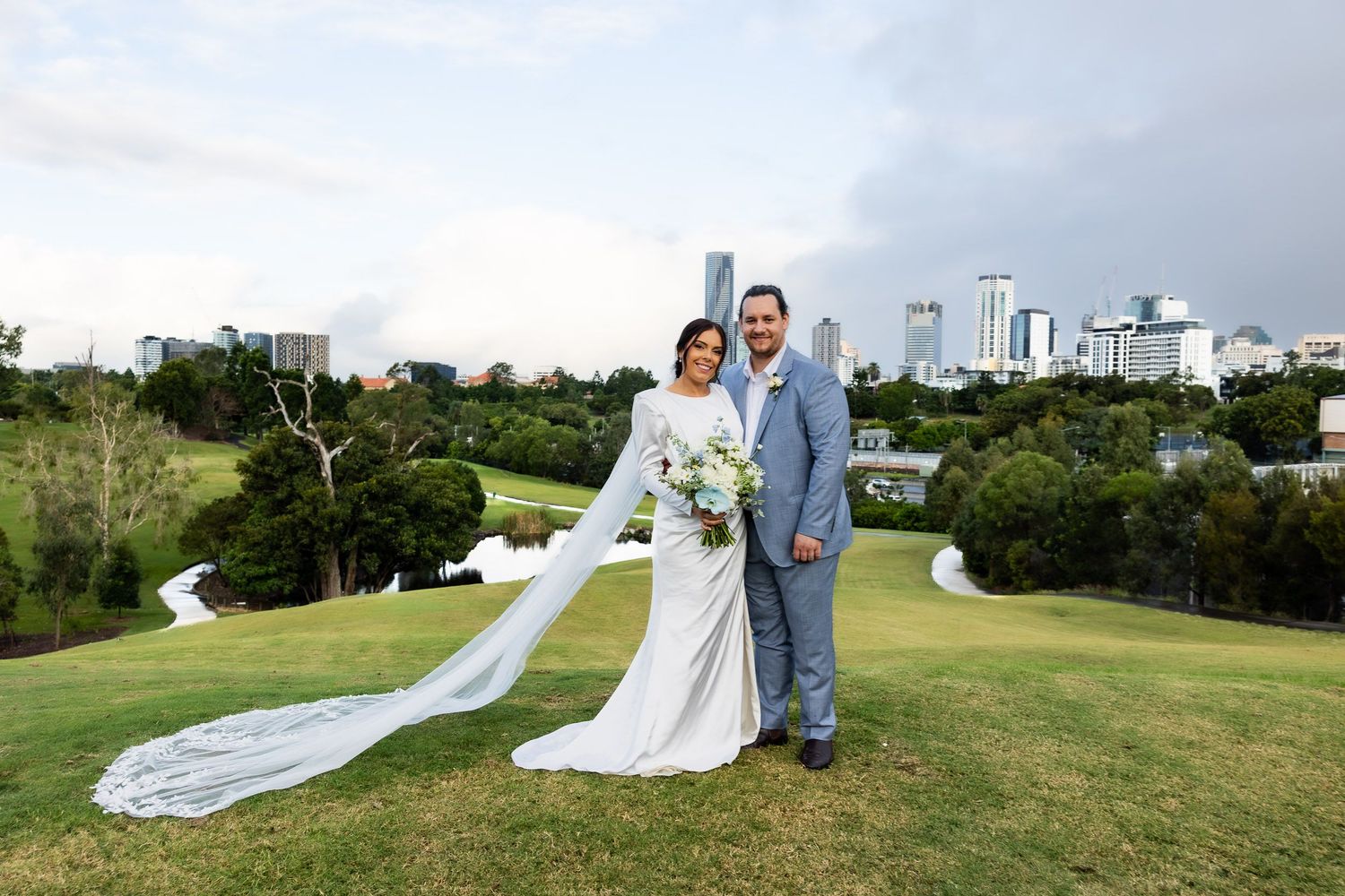 Wedding couple stands together on a green hillside with trees and buildings in the background on their special day.