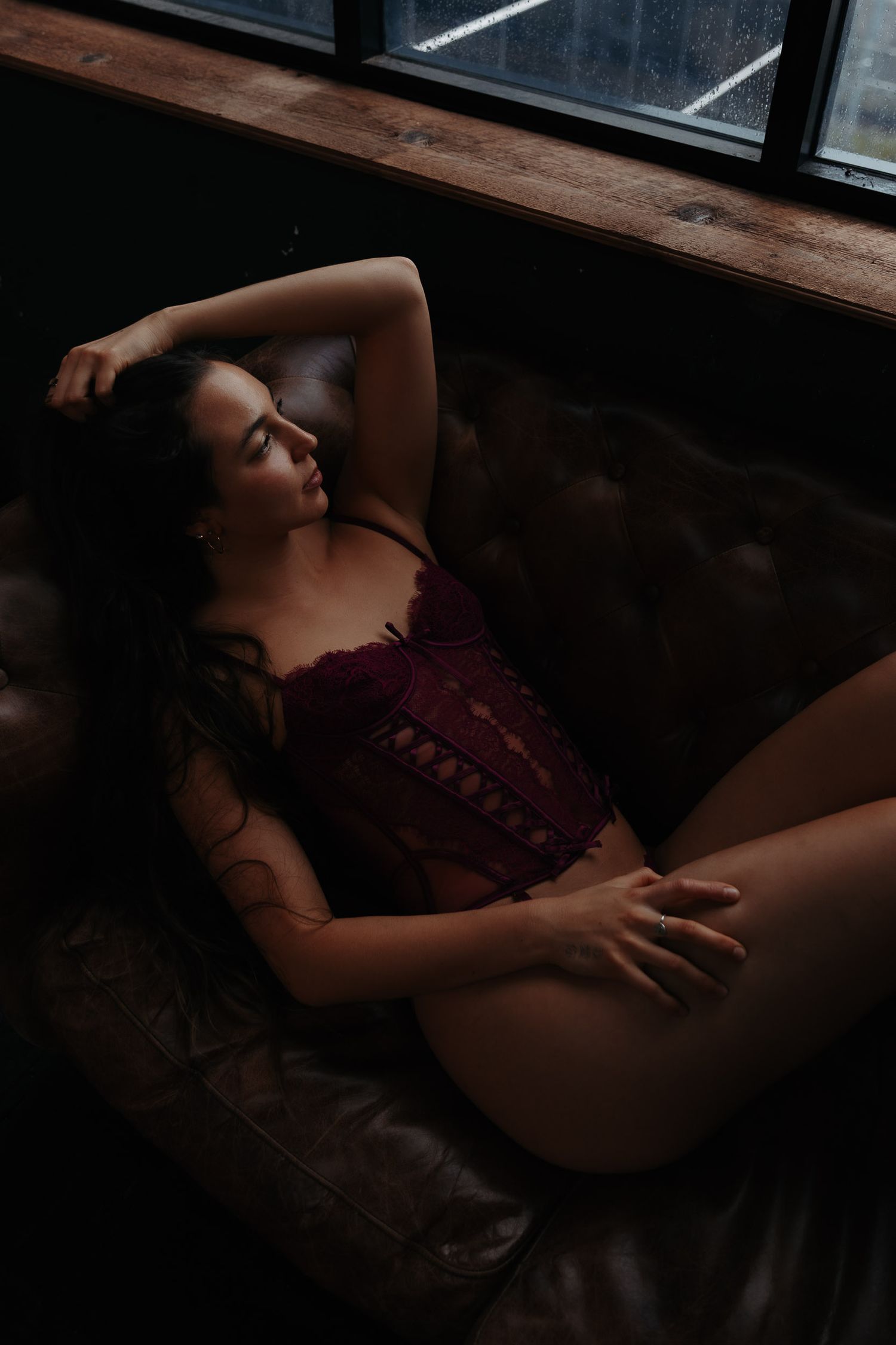 A person in burgundy lingerie lounges on a leather couch near a window in moody, dramatic lighting.