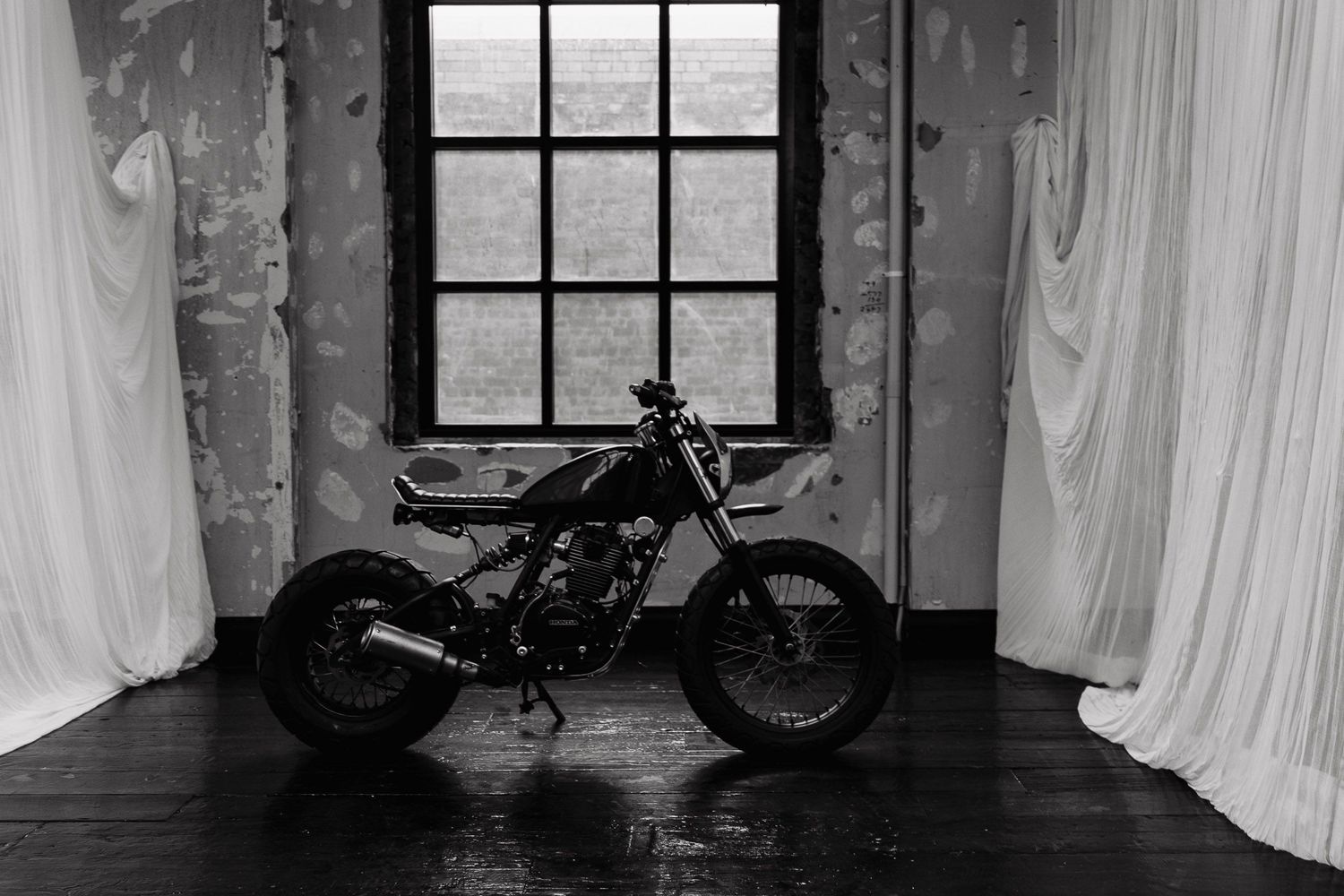 A black motorcycle silhouetted against a window with white curtains in a dark moody room, shot in black and white.