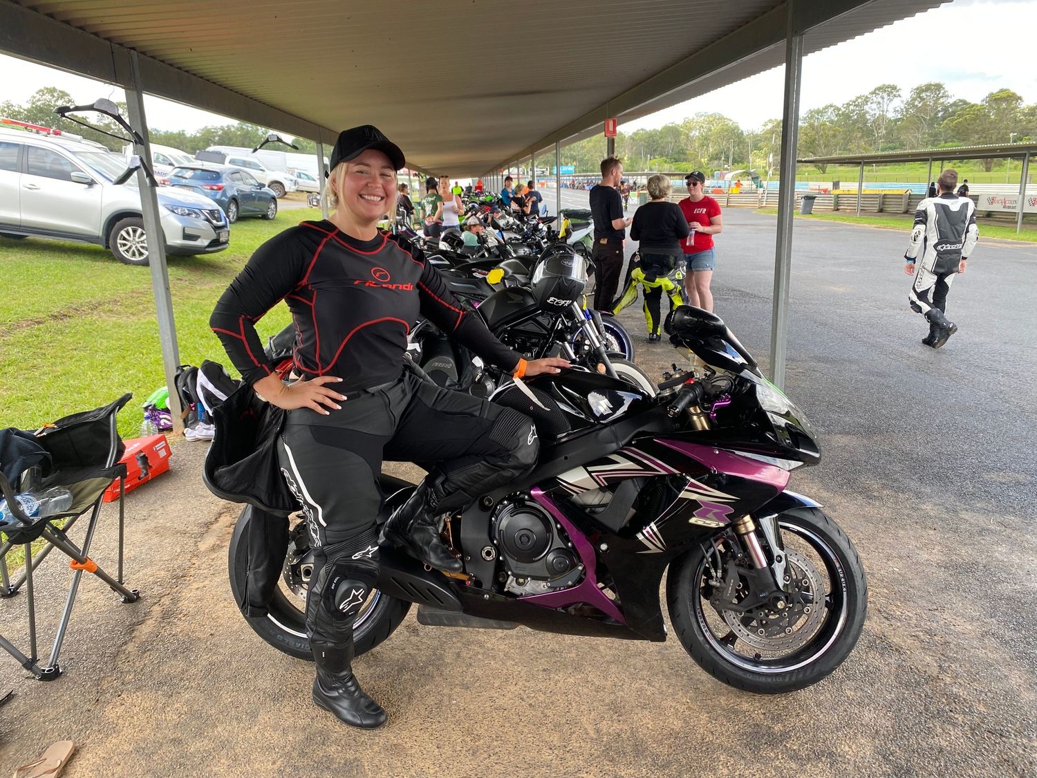 Motorcycles parked in line under a covered area with riders gathering around them during a group meetup.