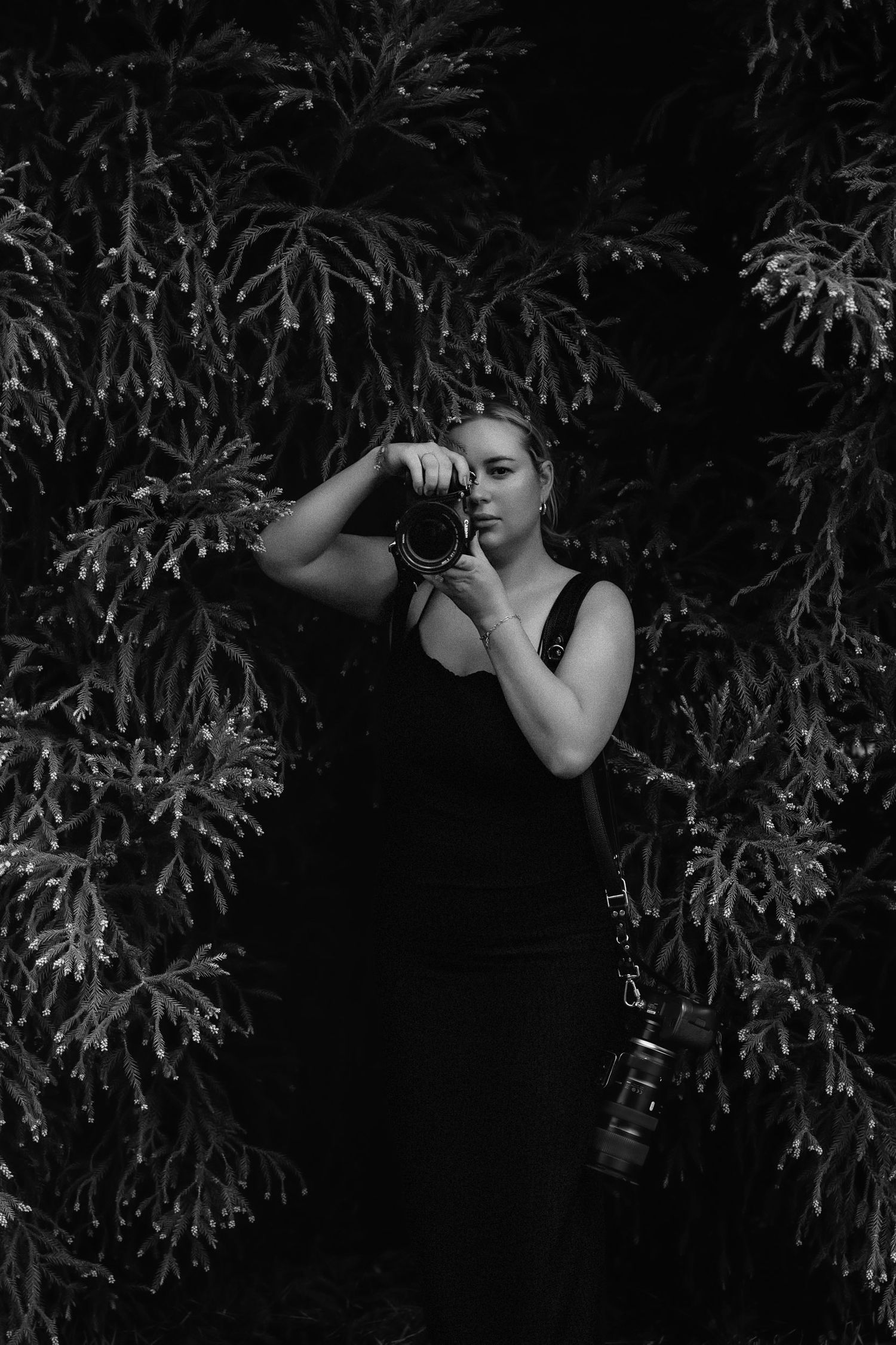 A photographer in a black dress takes photos among lush foliage in a moody black and white composition.