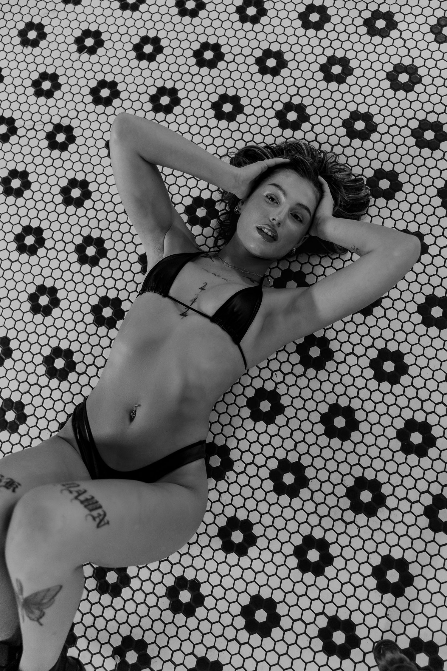 Black and white photograph of a figure reclining on a floral pattern tile floor in black swimwear.