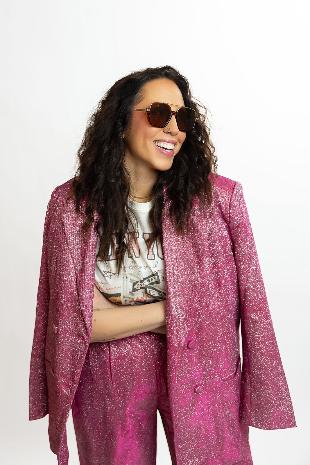 A person wearing a sparkly pink suit and sunglasses poses with a bright smile against a white background.