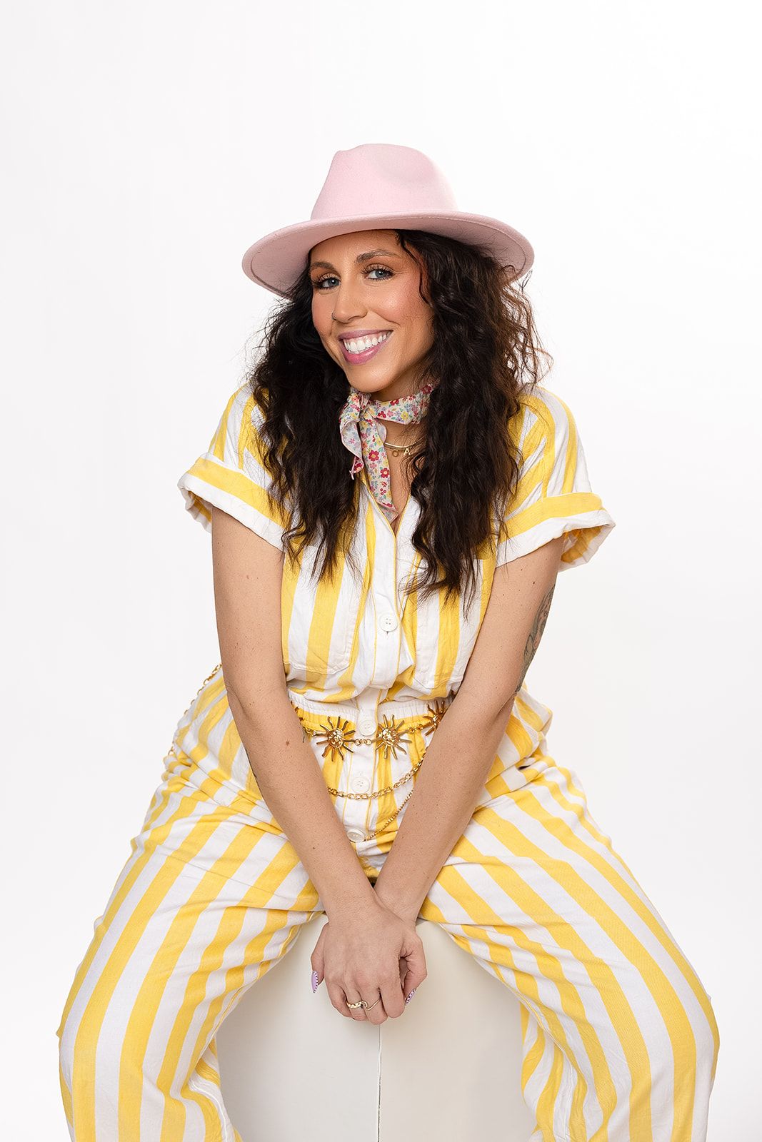 Smiling woman in yellow striped outfit and pink fedora hat seated against white background.