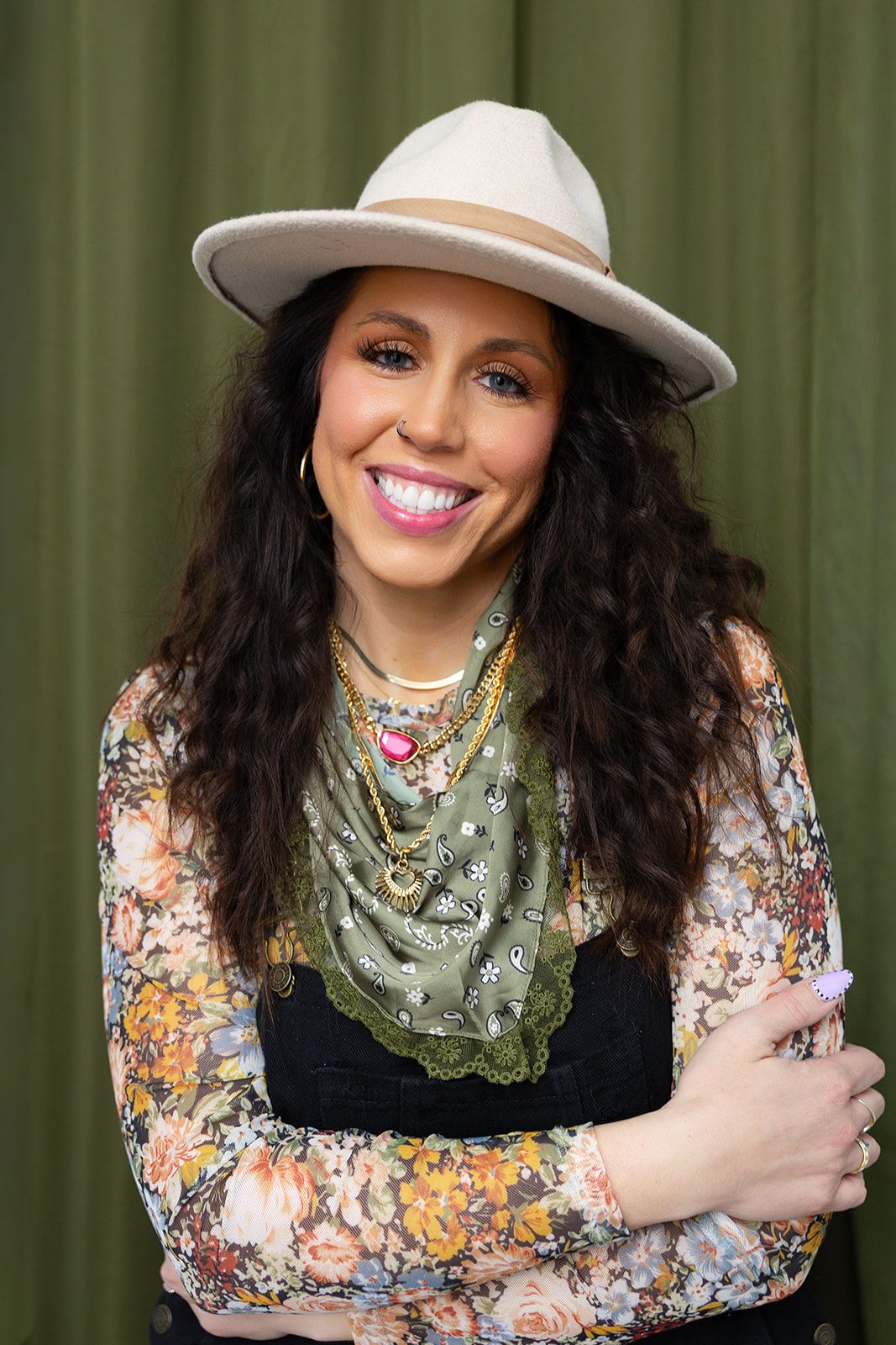 A person wearing a floral print top, white hat and sage green scarf poses against a green background.