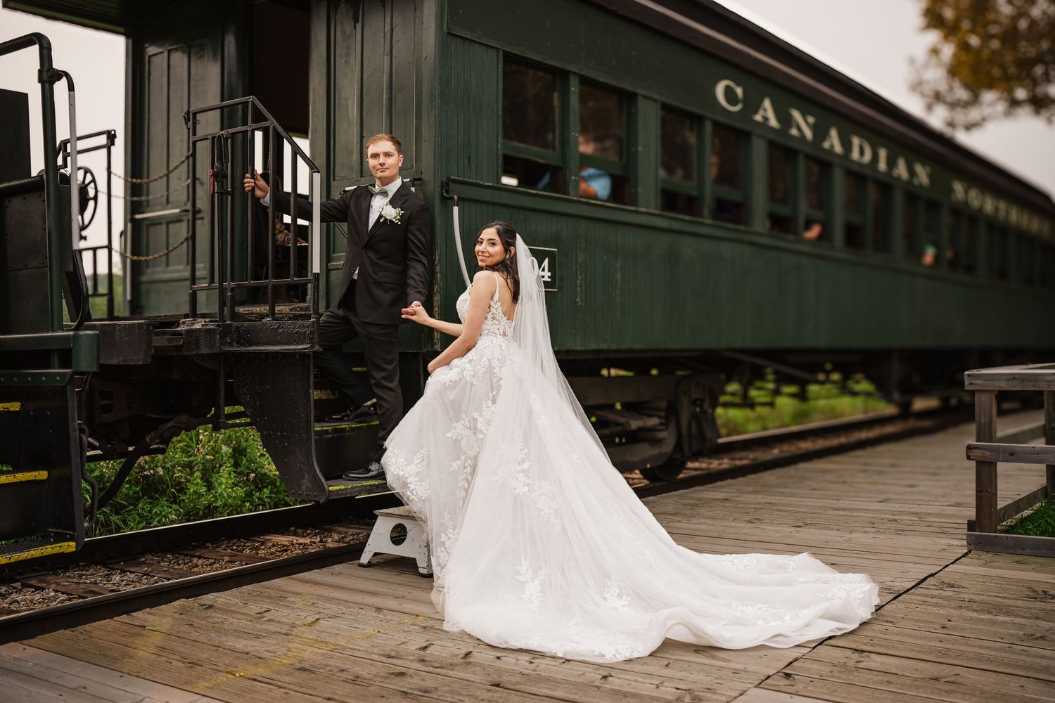 Bride in flowing white gown and groom in tuxedo pose beside a vintage Canadian Pacific railway train car at Fort Edmonton Park.
