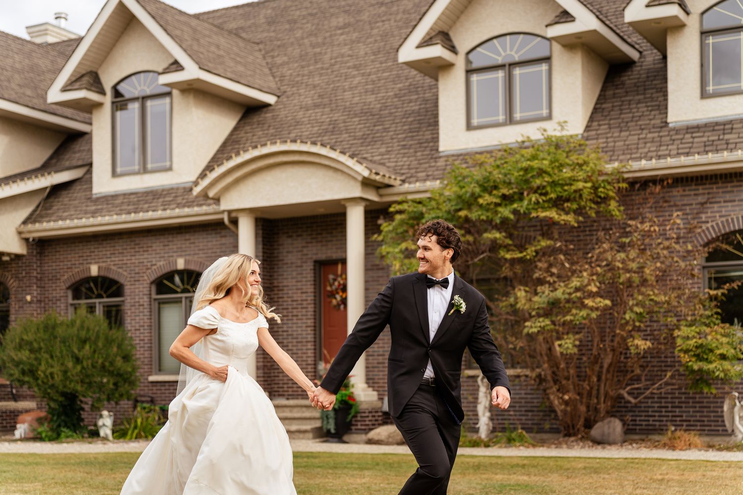 Bride and groom holding hands running joyfully in front of elegant brick mansion on their wedding day Cornerstone Wedding Venue.