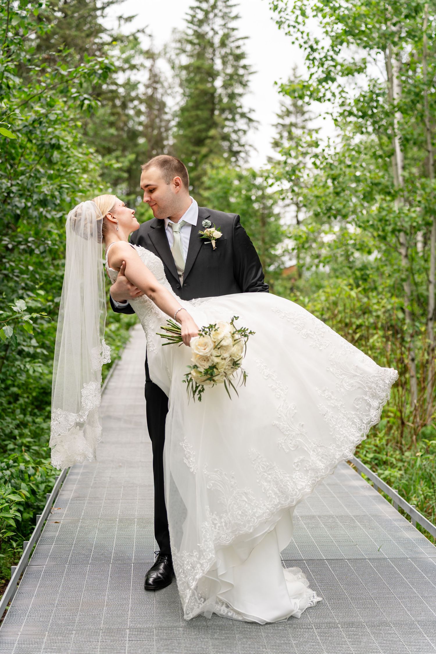Groom lifts bride in white lace gown on forest boardwalk, surrounded by lush green trees on their wedding day.