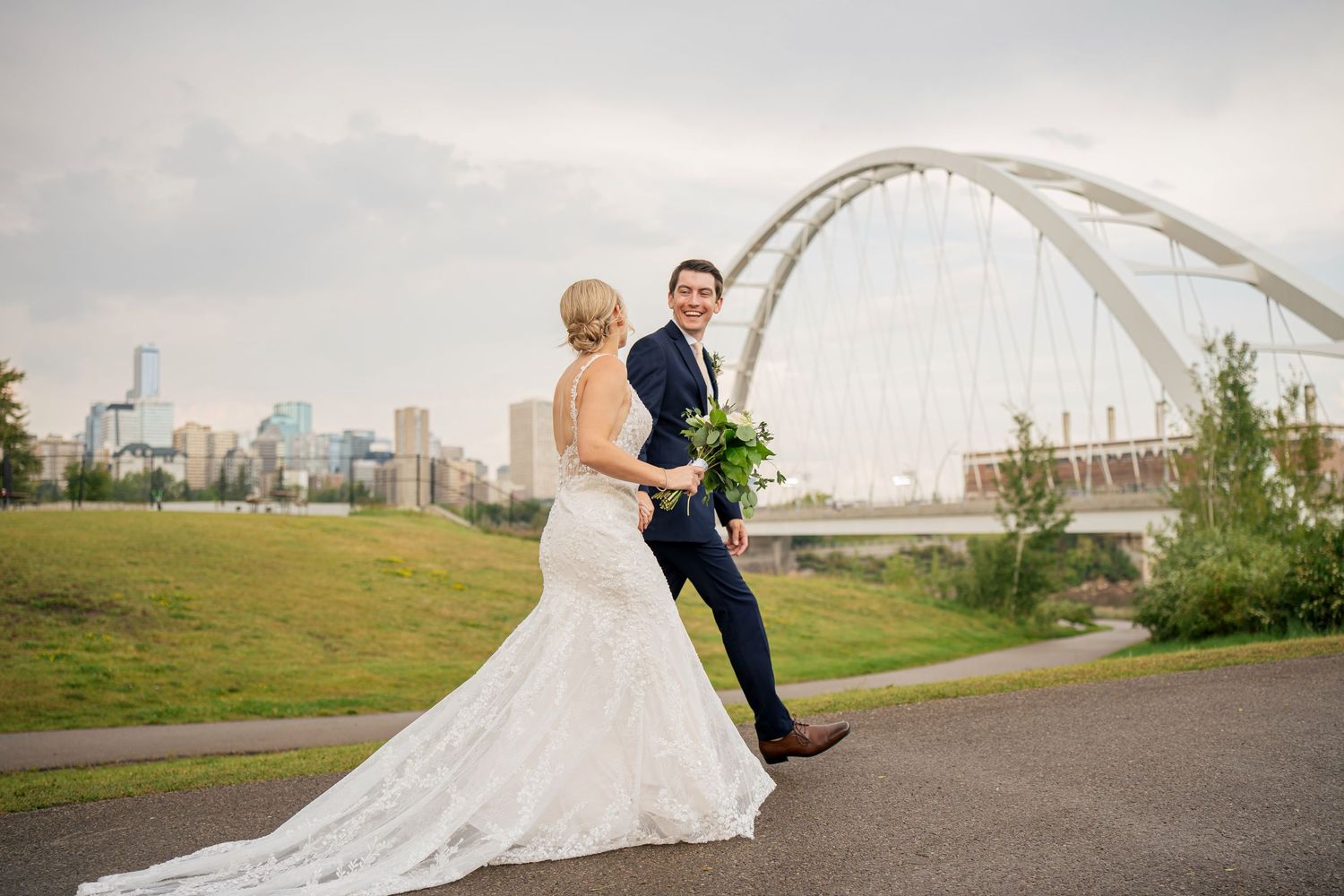 Bride and groom pose on a path with the Walterdale Bridge and Edmonton skyline in the background on their wedding day.