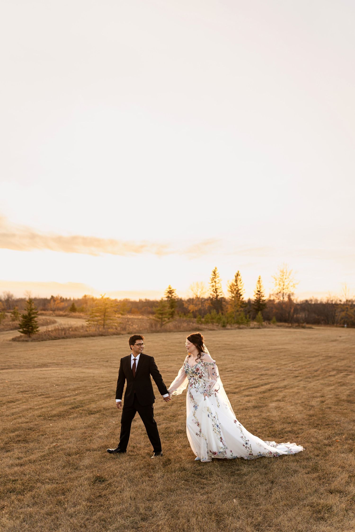 Bride and groom hold hands walking through golden field at sunset, bride wearing floral white gown.