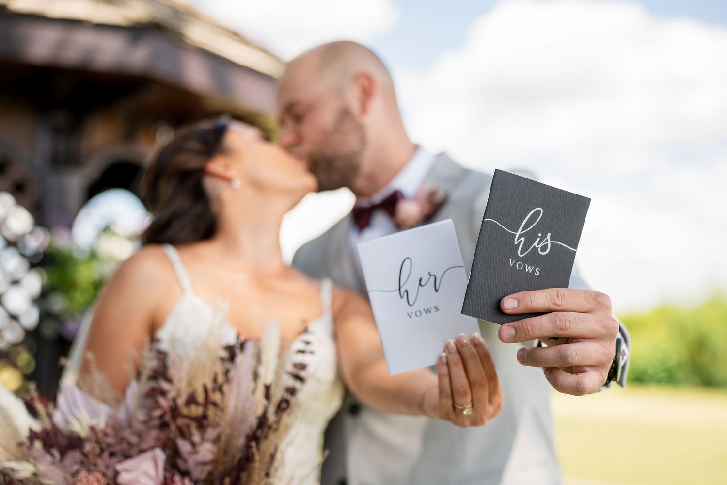 Newlyweds kissing while holding his and hers vow books outdoors at a wedding ceremony.