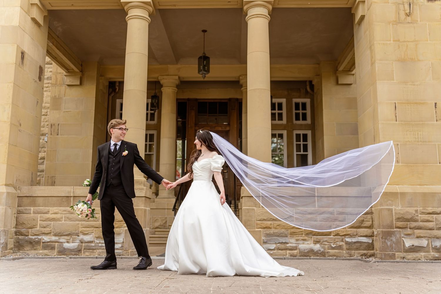 Bride and groom hold hands outside elegant stone mansion, her veil flowing in the breeze, bouquet in hand.