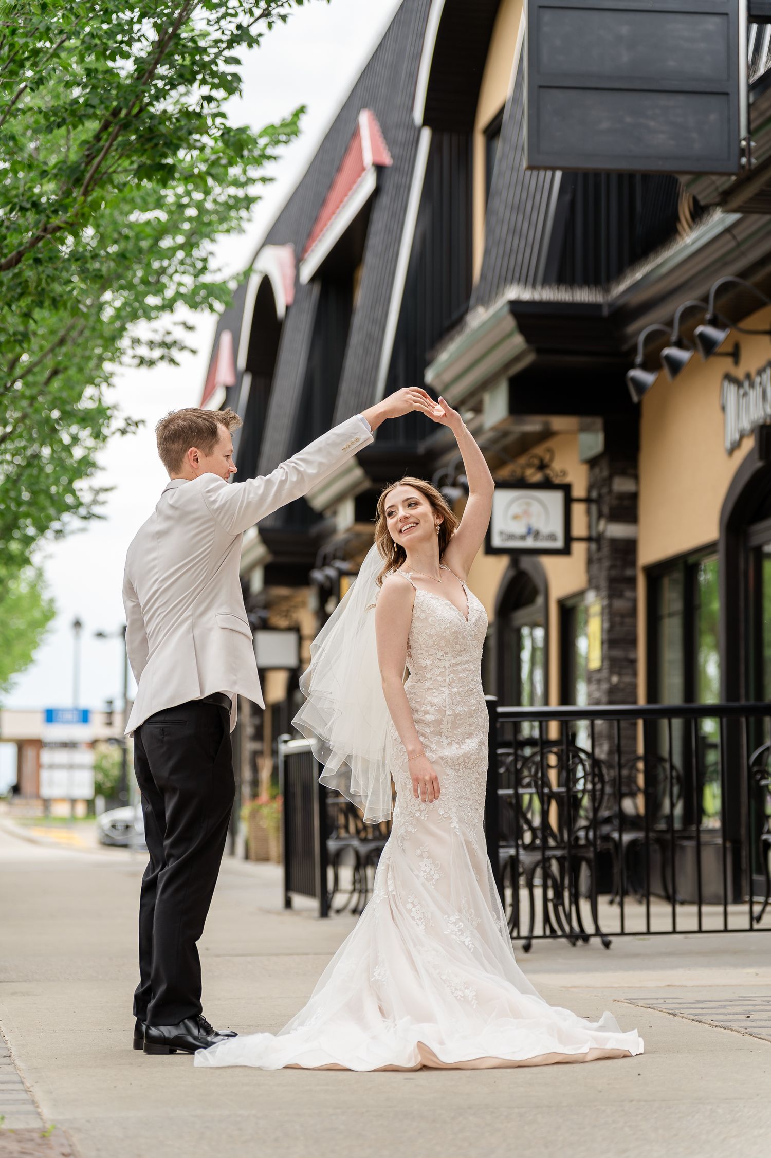 Groom twirling bride in white lace wedding gown on a charming outdoor street, sharing a joyful dance near a restaurant.