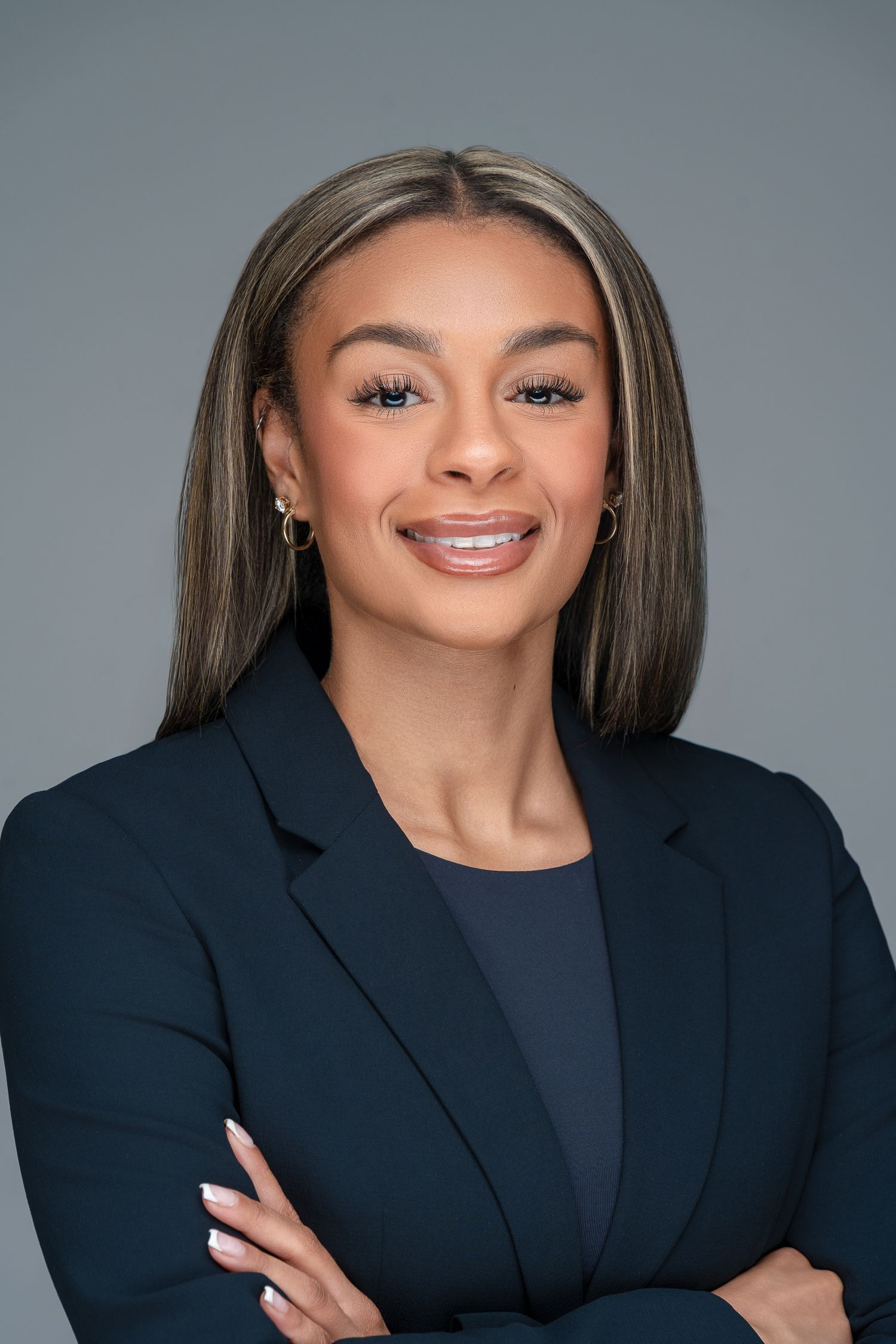 Professional headshot of a confident professional in a navy blazer posing with arms crossed against gray background.