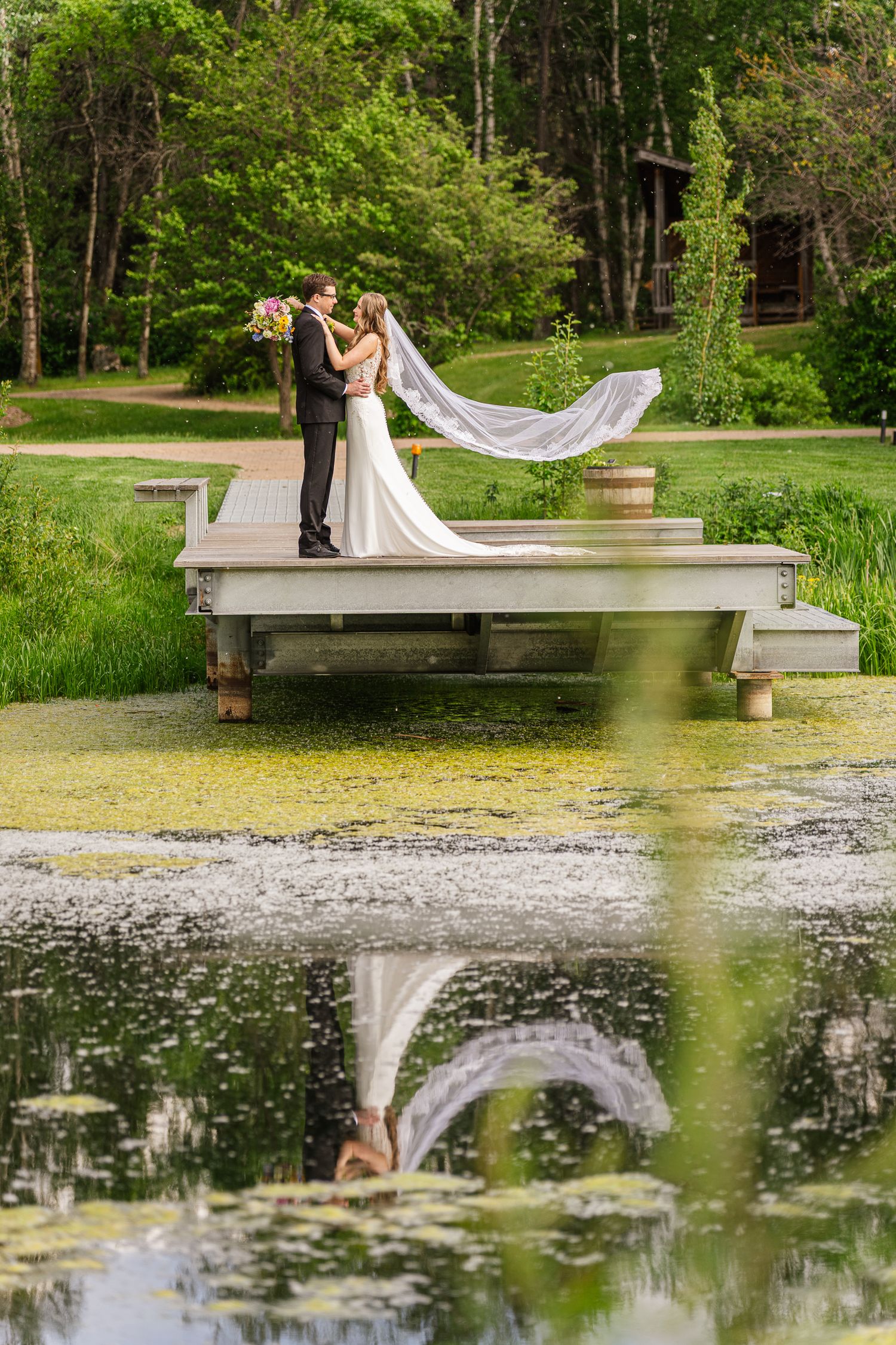 Bride and groom kissing on a dock over a lily pond, with veil flowing, reflected in still water, surrounded by lush greenery.