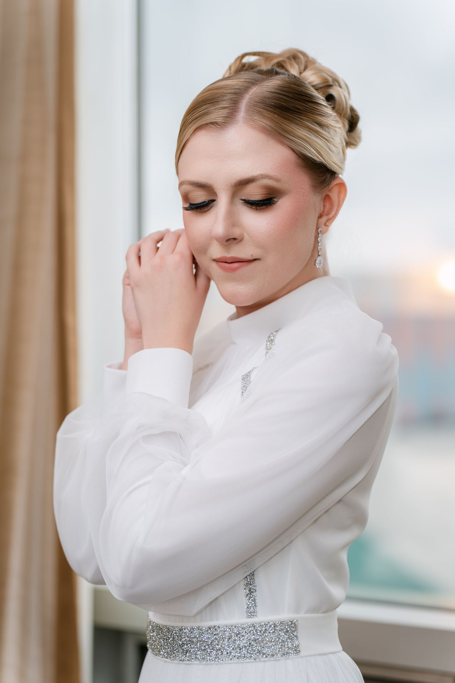 Bride in elegant high-neck white wedding dress with crystal belt poses gracefully near window in series of photos.