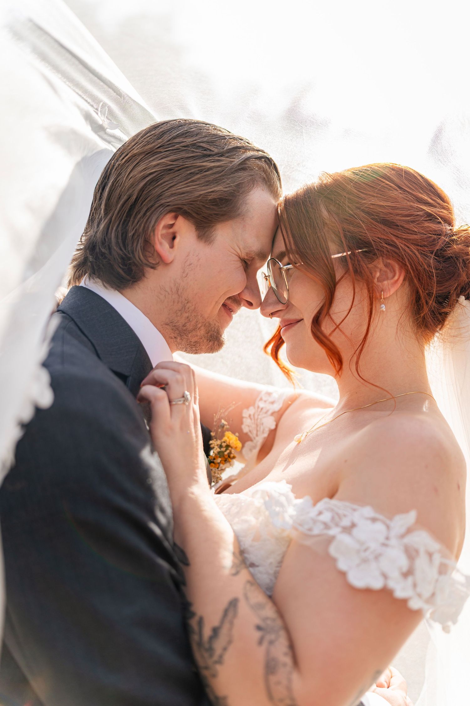 Romantic wedding couple embraces in soft sunlight with bride wearing lace off-shoulder dress and groom in navy suit.