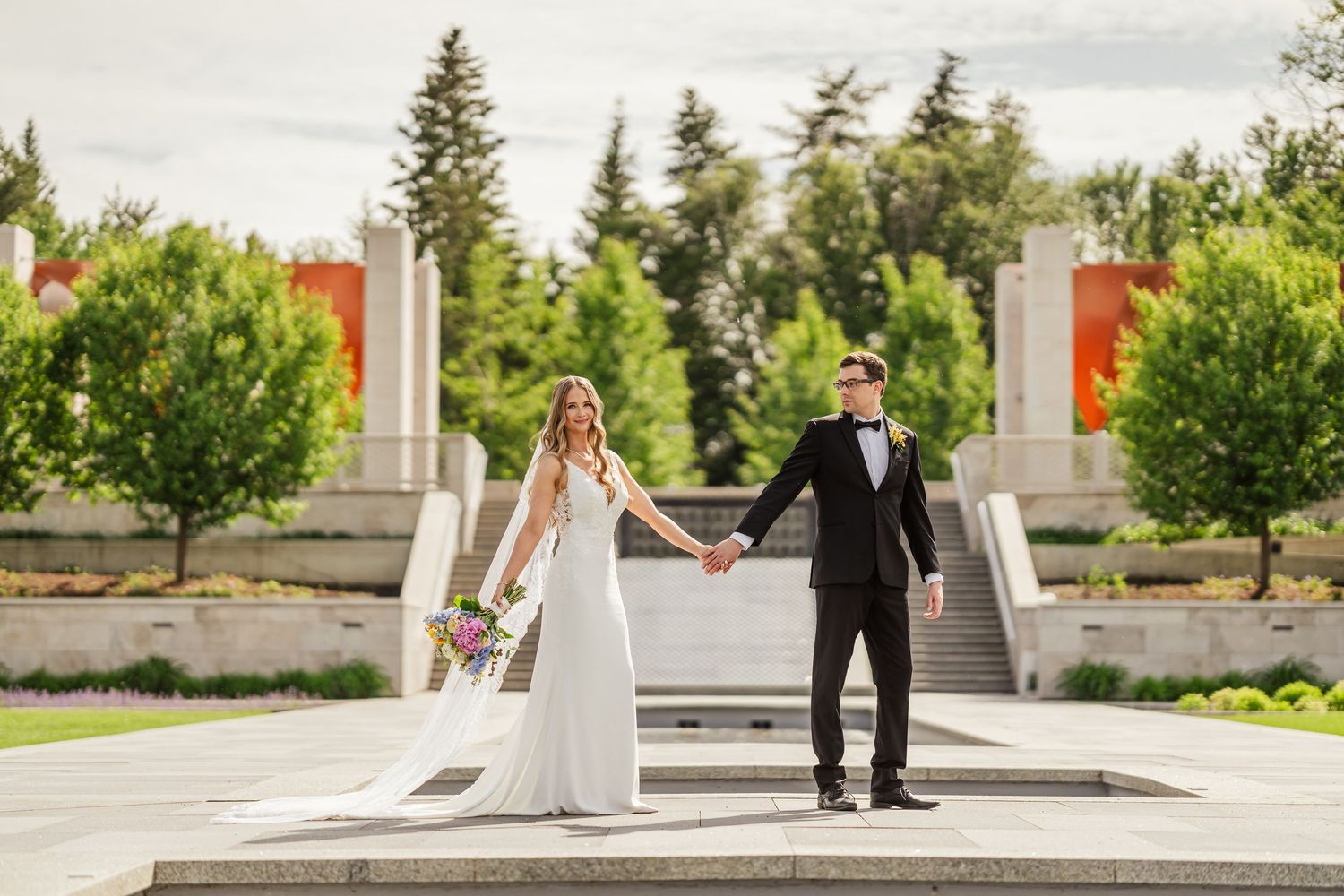 Bride and groom holding hands outdoors at a modern garden venue with lush green trees and architectural columns At U of A Botanic Garden.