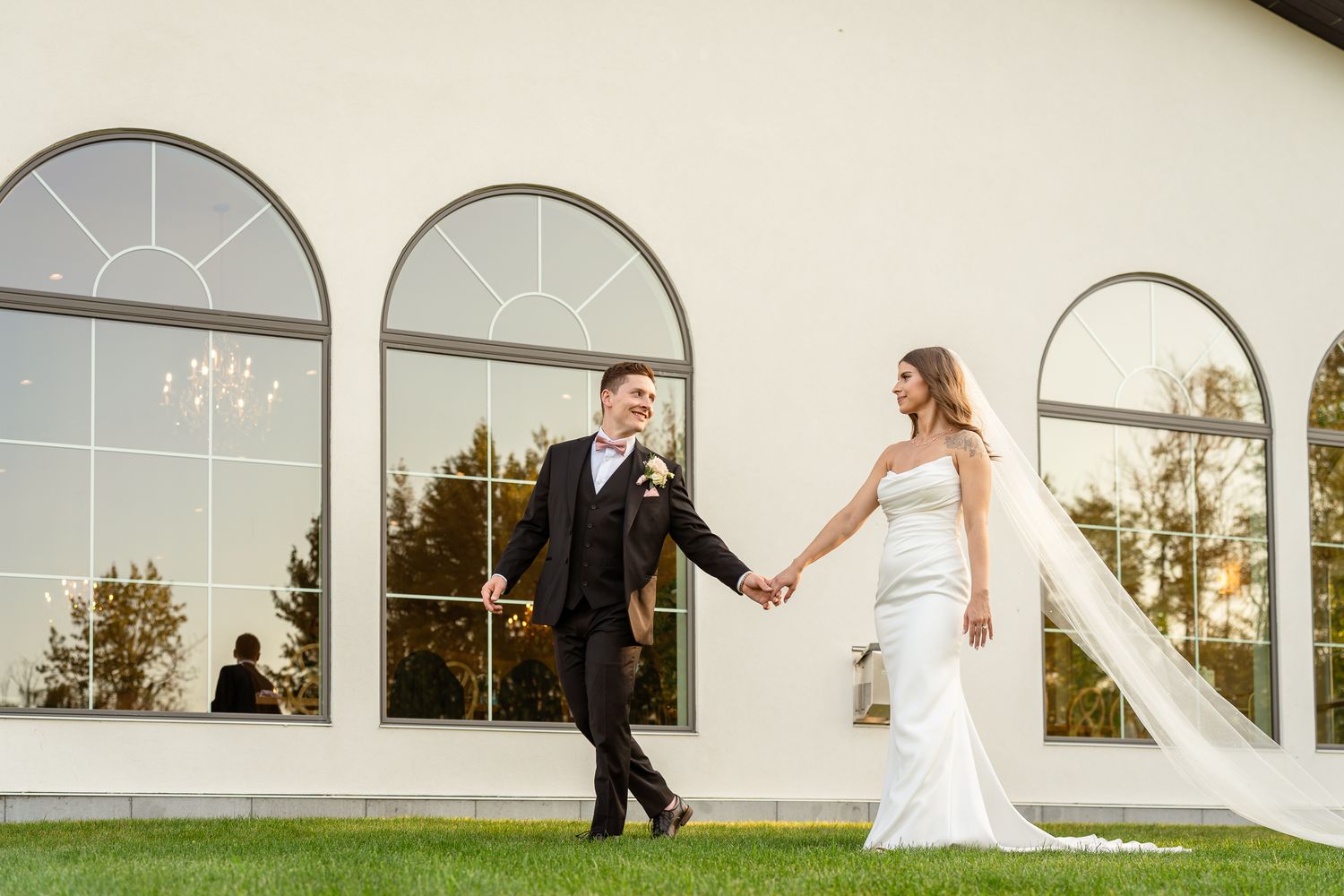 Bride and groom holding hands walking on green grass in front of elegant white building with arched windows Sparrow Lane Events.
