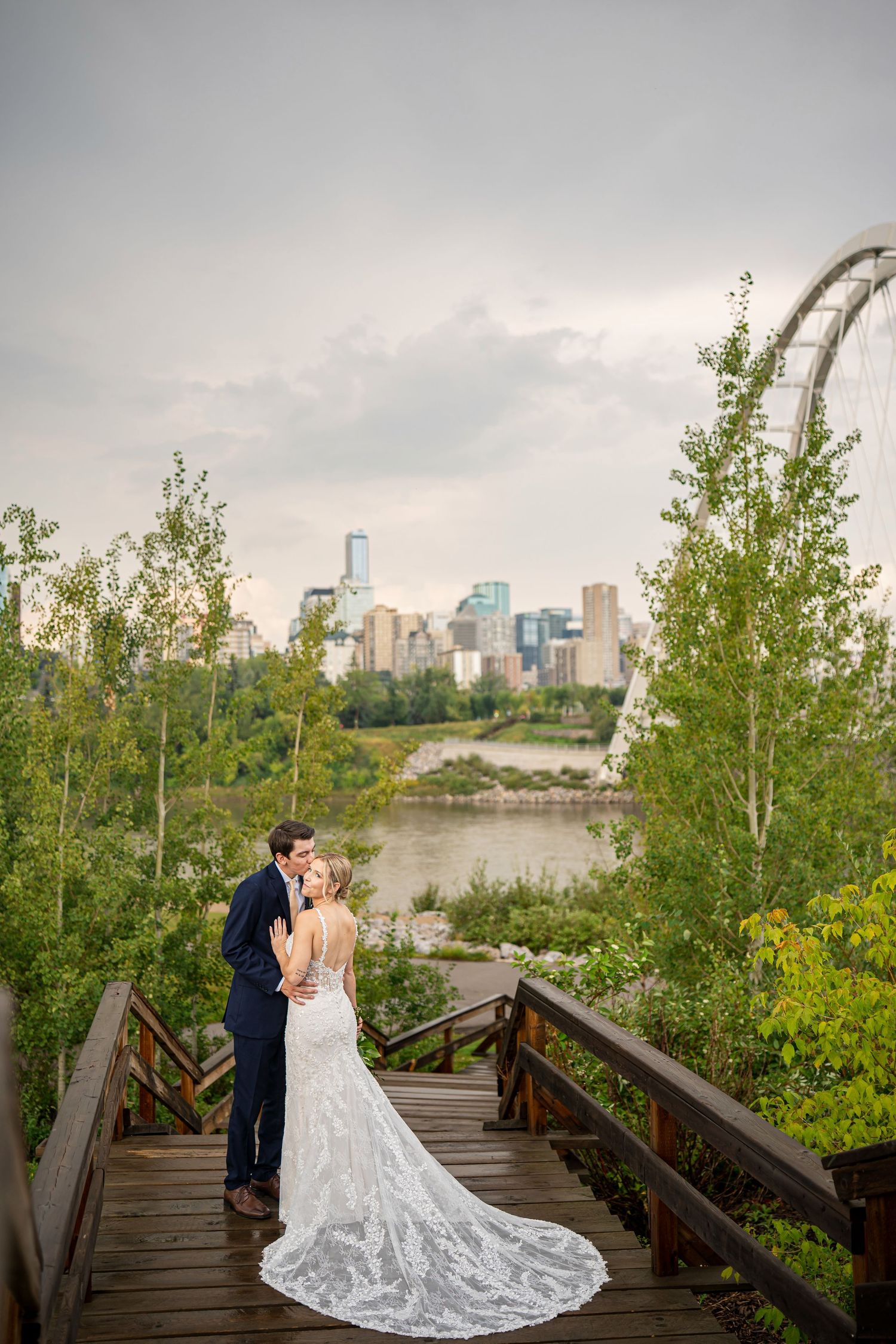Bride and groom embrace on wooden steps with lush greenery and city skyline backdrop on their wedding day.