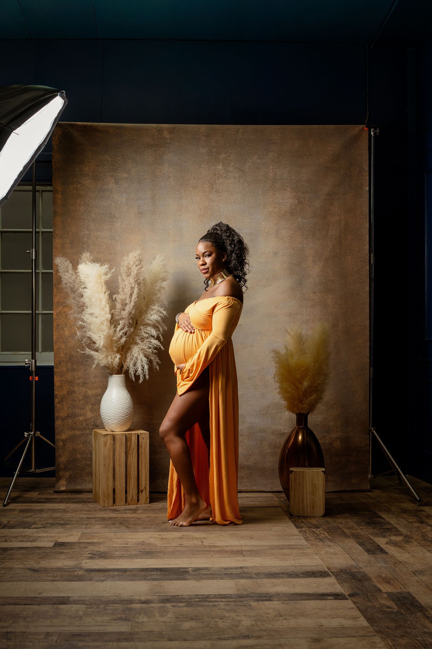 A maternity photoshoot featuring a yellow-orange flowing dress against a brown backdrop with pampas grass decor in a studio.